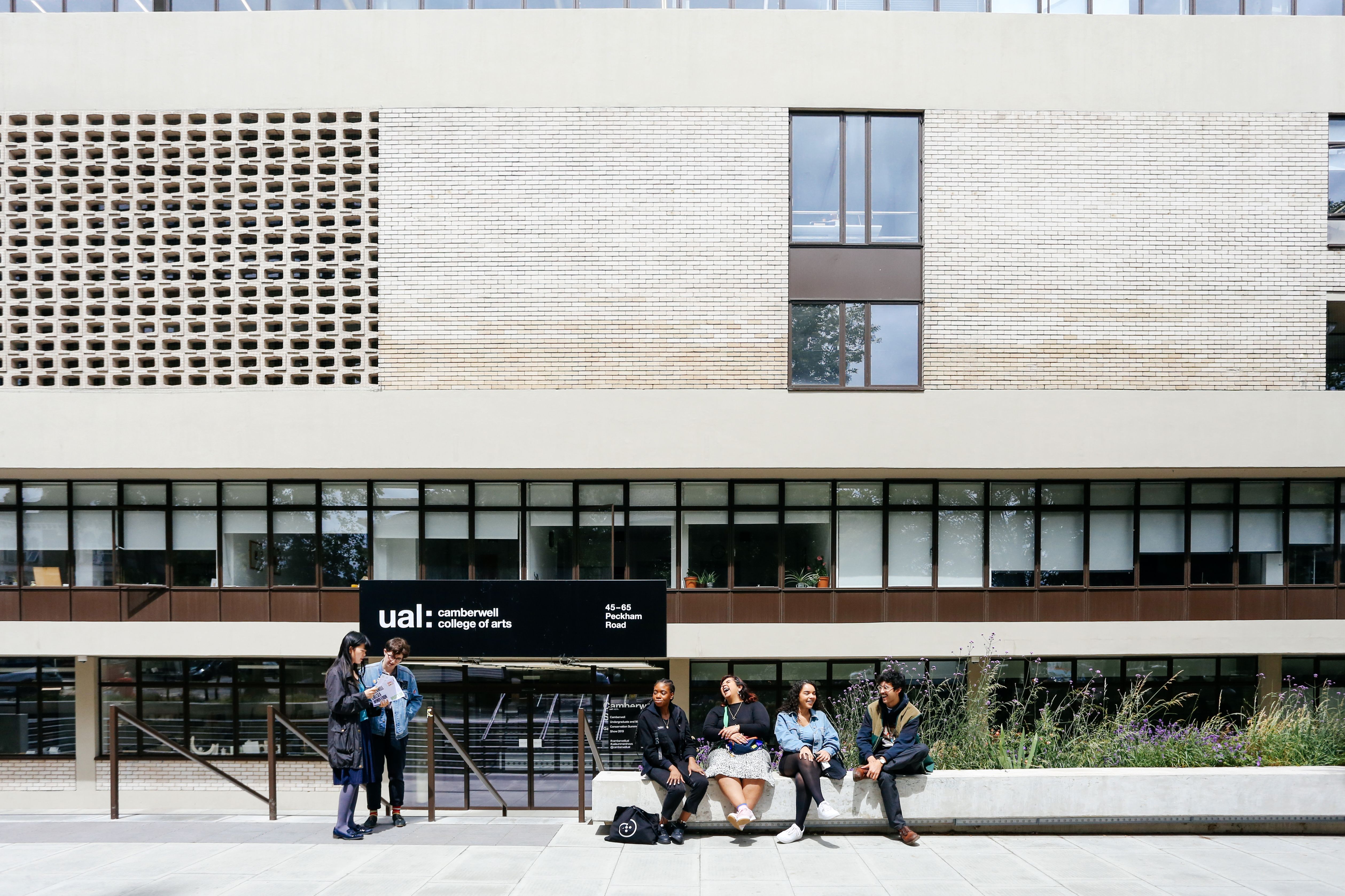 Students sitting outside of a large building with the words 'UAL Camberwell College of Arts' on the front of it. 