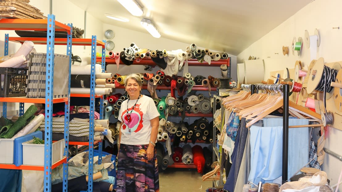 A white woman with short hair is standing in a store with lots of fabric rolls on shelves behind her and on each side. She is wearing a t-shirt with a printed pattern, a necklace and a skirt.