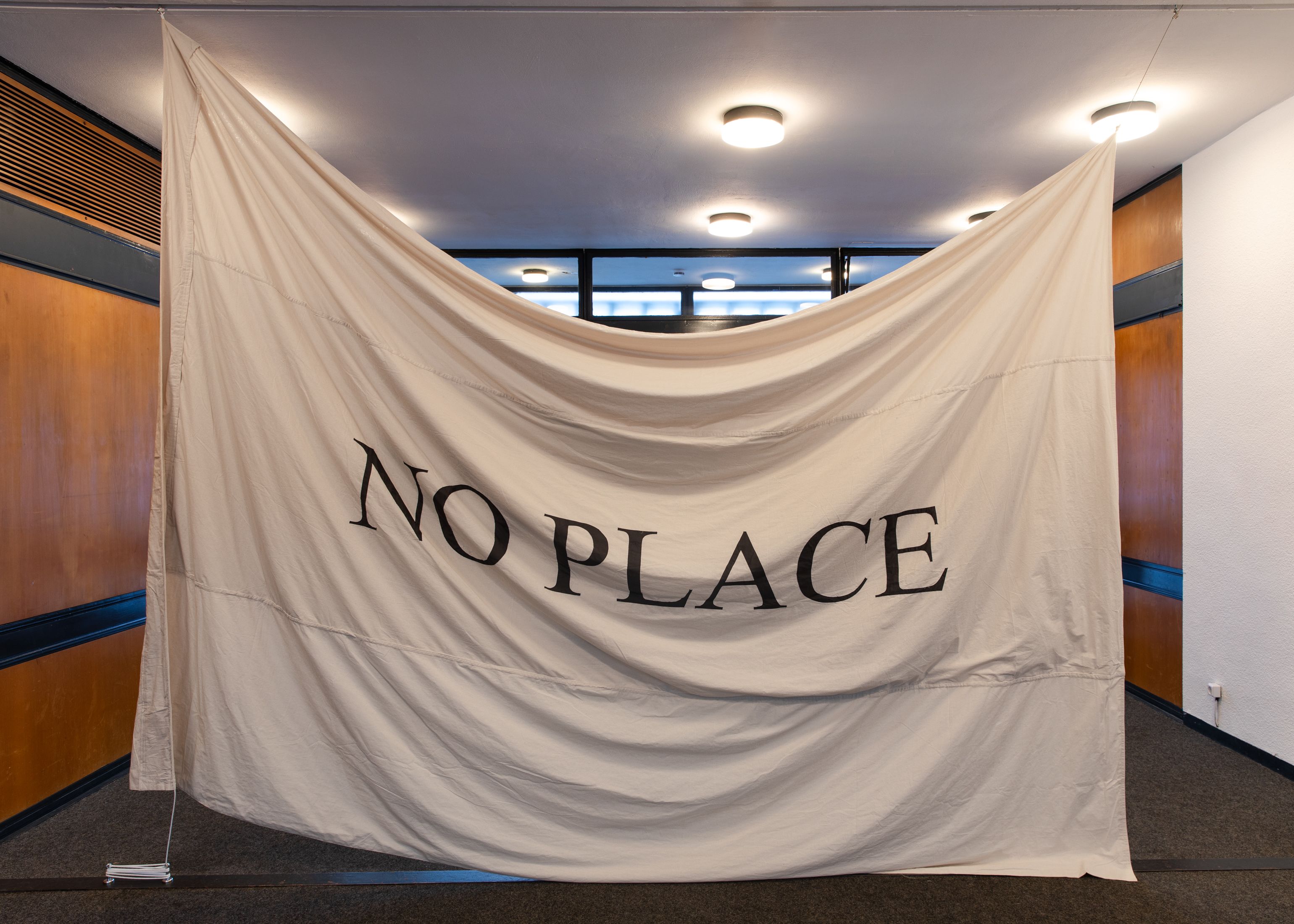Large piece of material strung from the ceiling with 'NO PLACE' printed in a large black font in the middle.