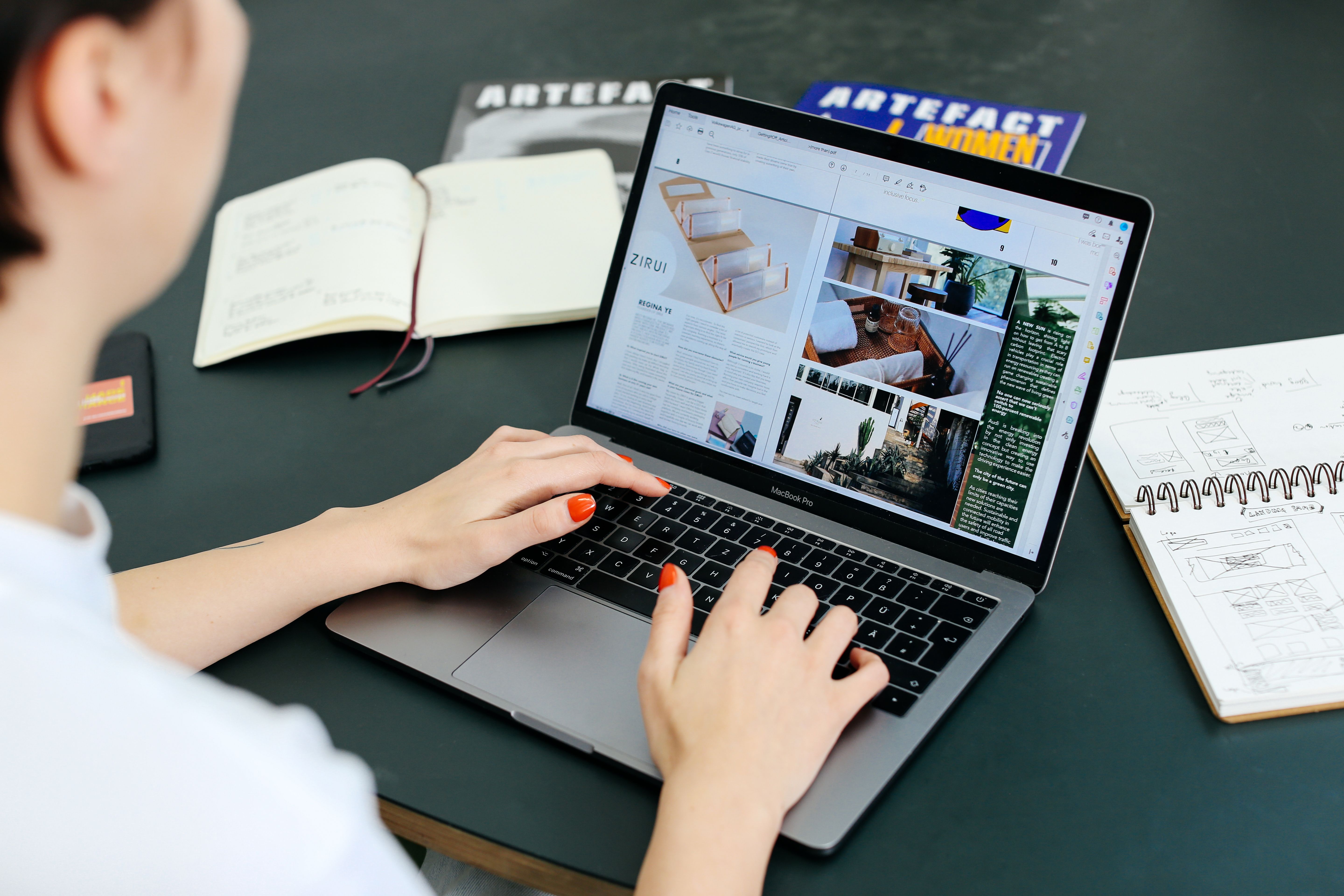 Person typing on laptops with 2 notebooks and an open magazine on the table.