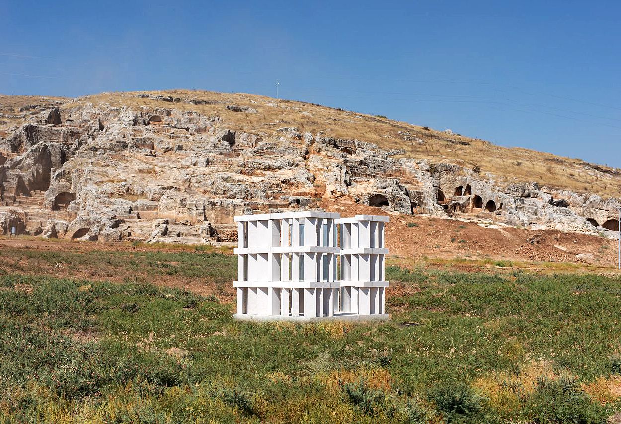Rugged rural landscape with a white sculpture in the middle. The sculpture is a square made of a blocks, with spaces in-between. 