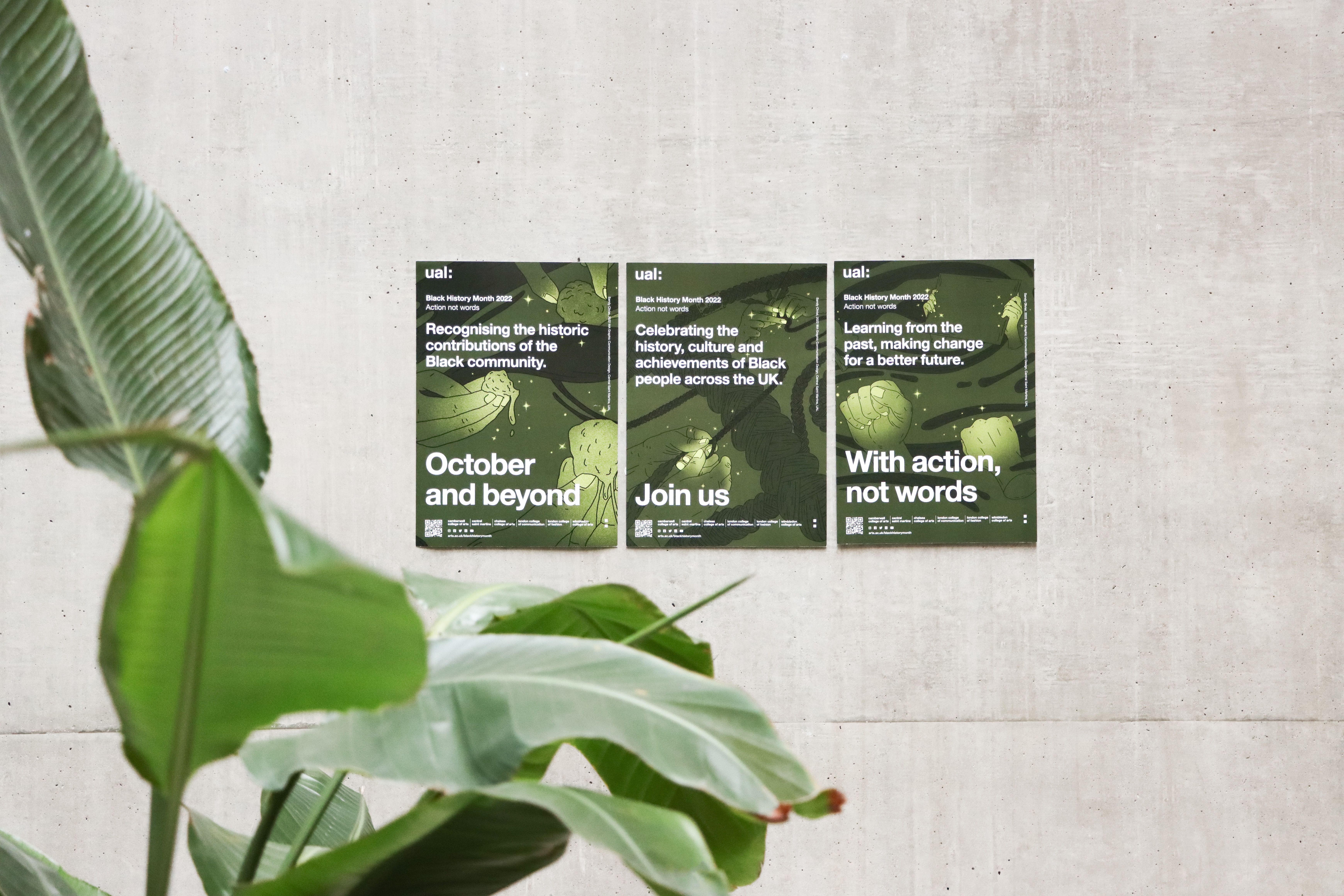Three posters displayed on a concrete wall promoting UAL Black History Month 2022. The posters highlight themes such as recognizing the contributions of the Black community, celebrating culture and achievements, and advocating for action over words. Large green leaves frame the scene in the foreground.