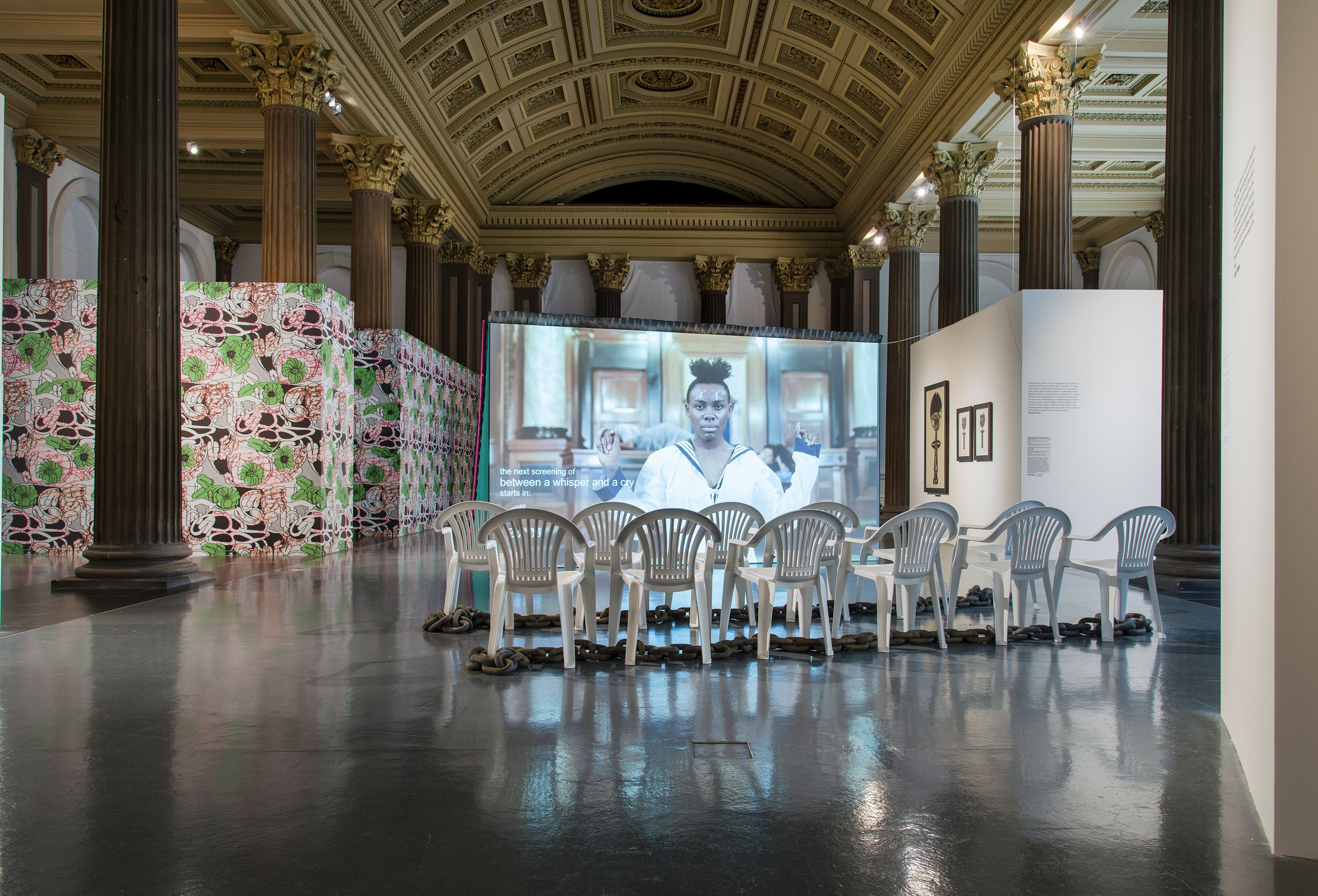 A museum with ornate ceiling and artworks on the walls and on screen. There is a reflective black floor with white garden chairs place in front of the screen. A large metal chain runs along the floor between the chairs