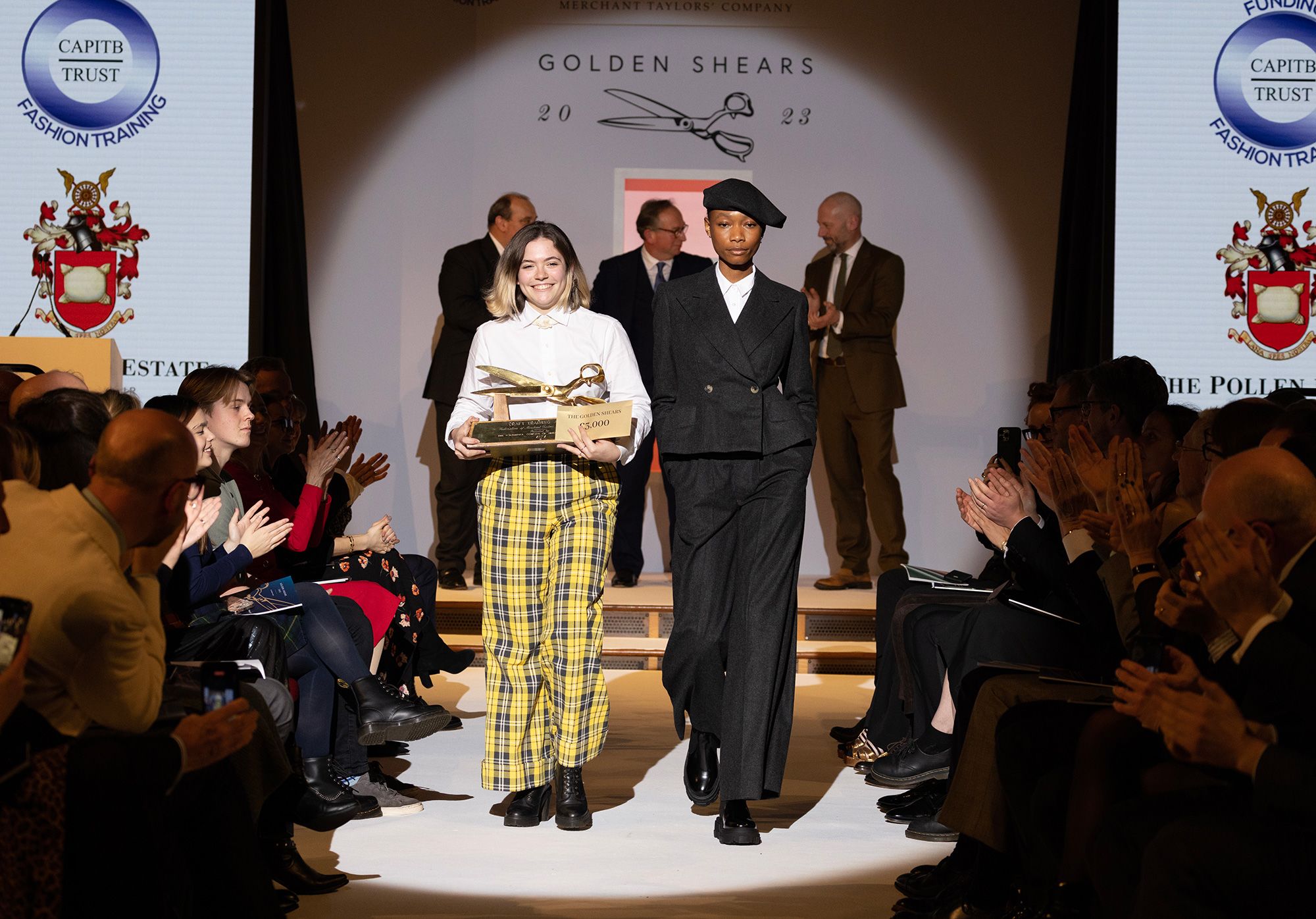 Image shows 2 people walking down a fashion runway with a spotlight on them and audience on either side. The person on the left is wearing checked yellow trousers and is holding an award. The person on the right is wearing a sleek black suit. The audience around them are clapping.