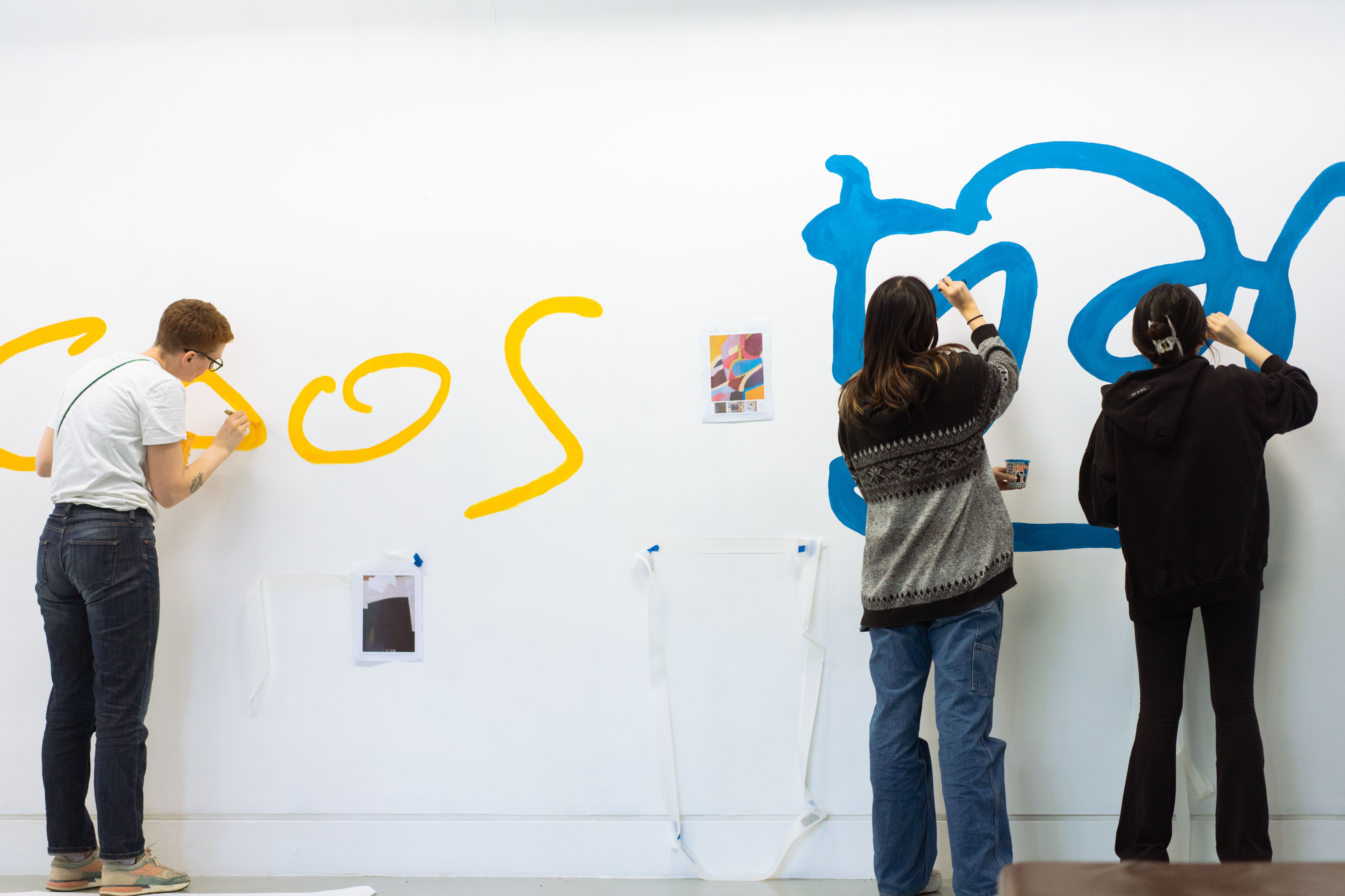 Students with their backs to camera painting large lettering in blue and yellow on a white wall