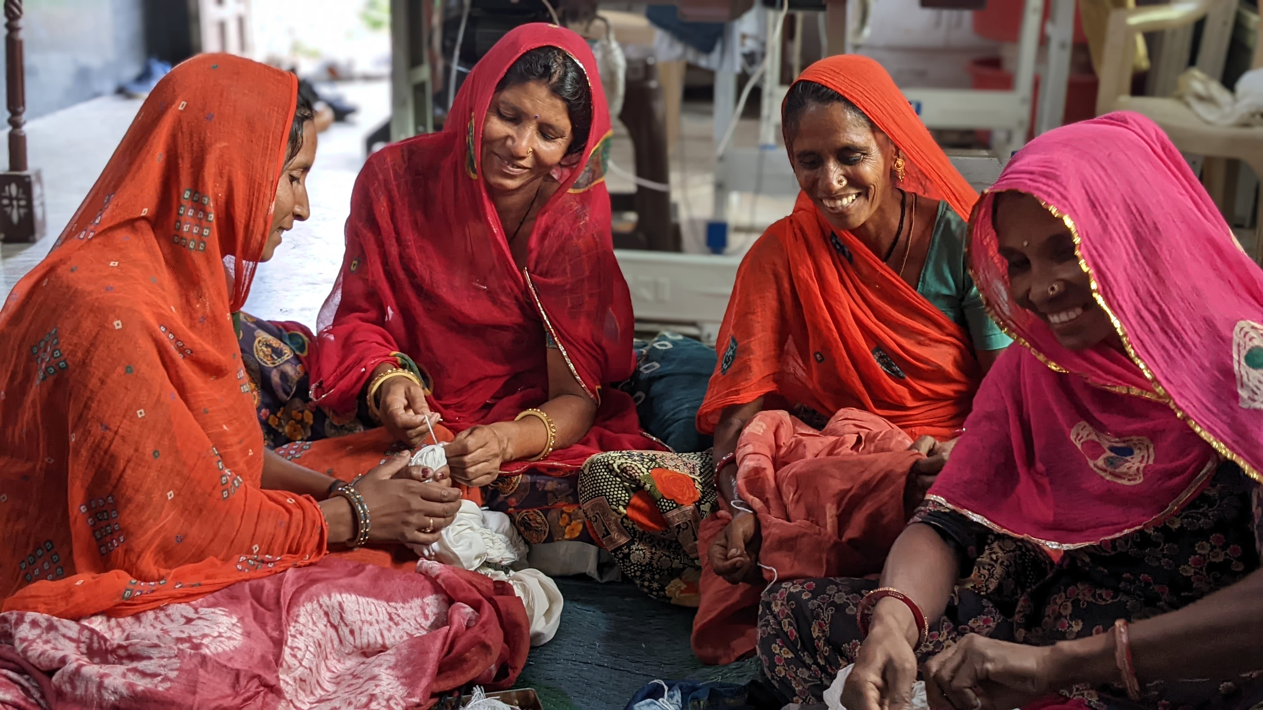 Four women sitting in a circle sewing. They're all wearing tones of red, orange and pink. They're smiling as they're working. 