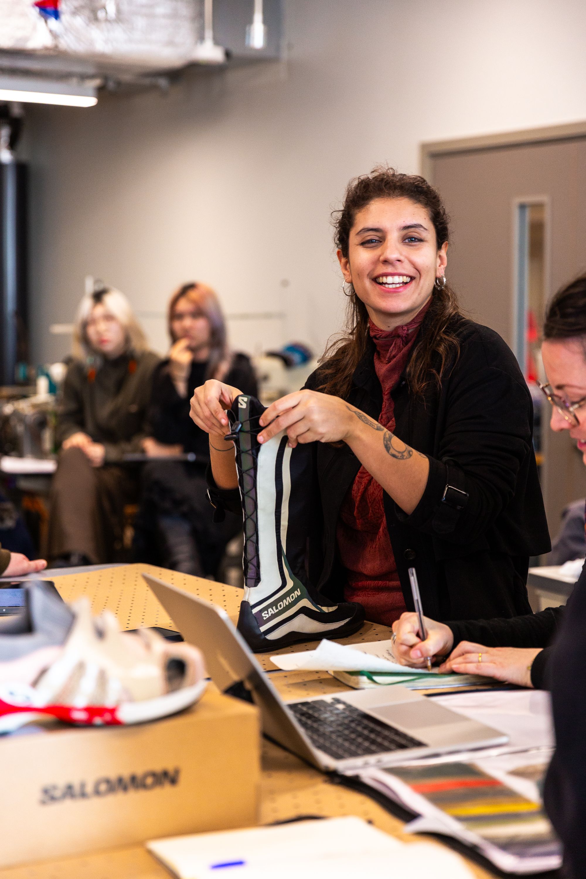 A woman holding a boot and smiling towards the camera with two people in the distant background and another person writing with a pen.