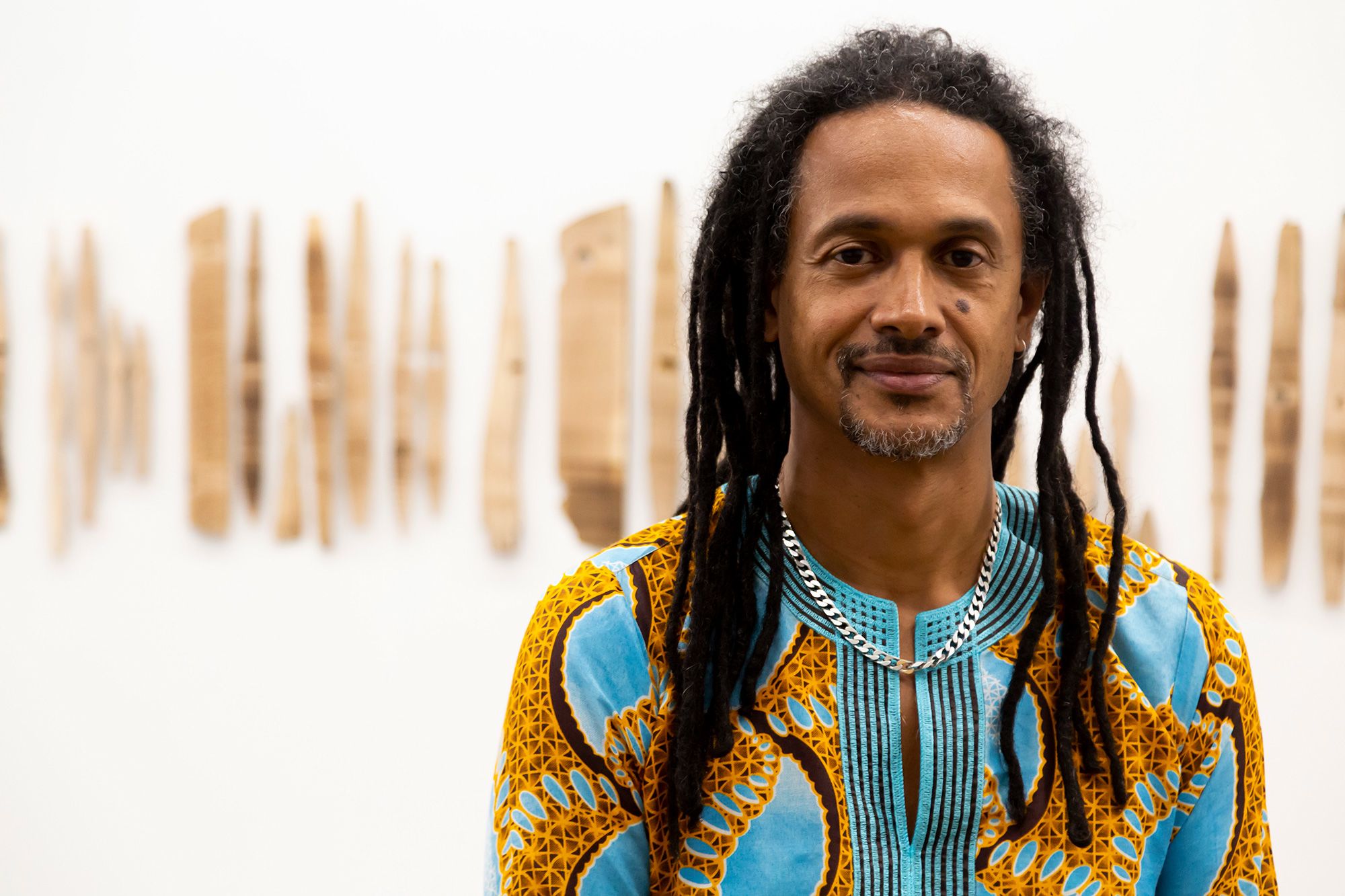 Image shows a man with long black dreadlocks wearing a colourful yellow and turquoise shirt standing smiling at the camera