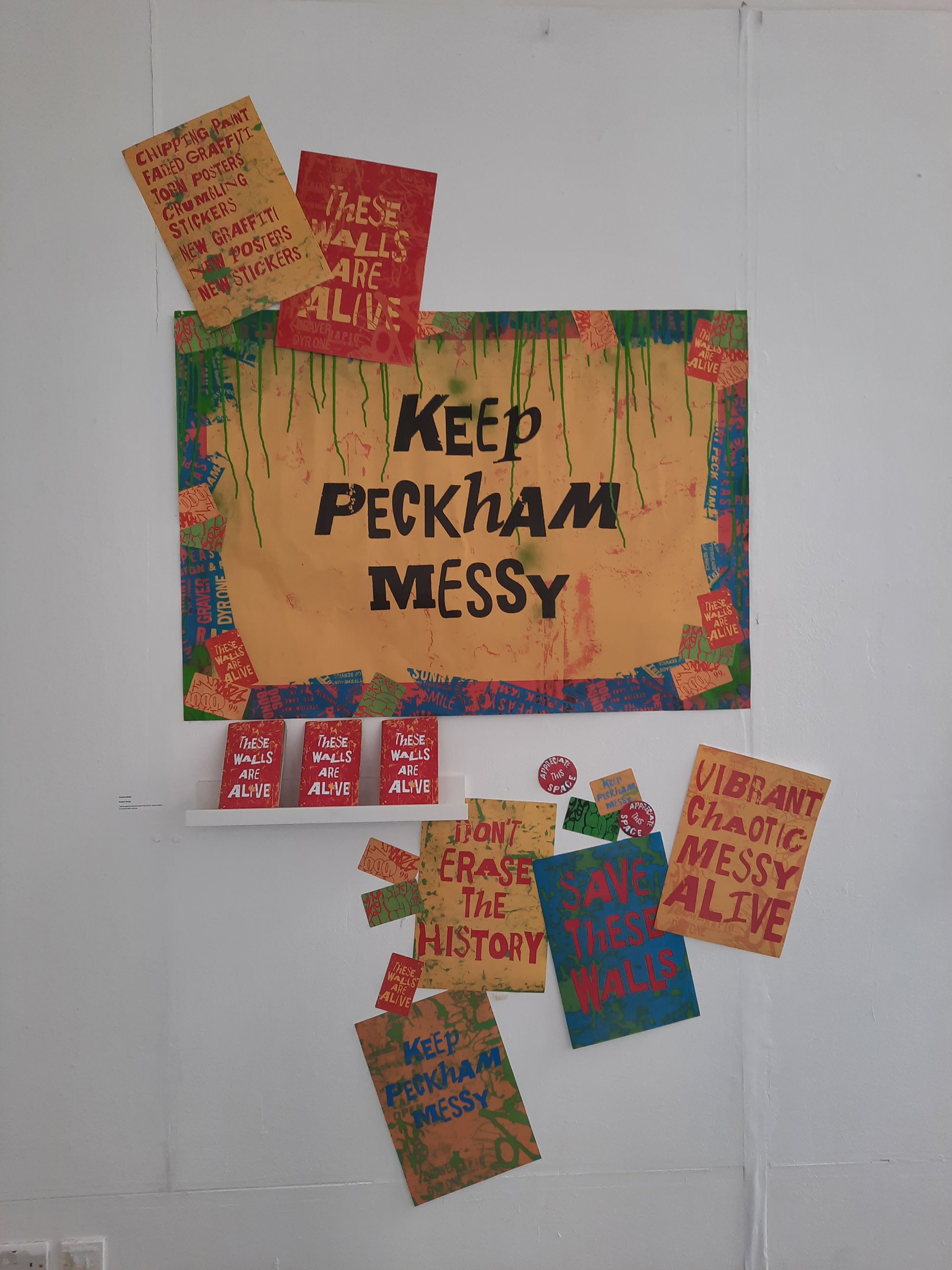 Image shows a large landscape poster with the words ‘ Keep Peckham Messy’ which is centred, the post is surrounded by smaller portrait posters, stickers and postcodes saying ‘ These walls are alive’ ‘Don’t erase the history’ and ‘Vibrant, Chaotic, Messy, Alive’.  Colours used are yellows, reds and greens.