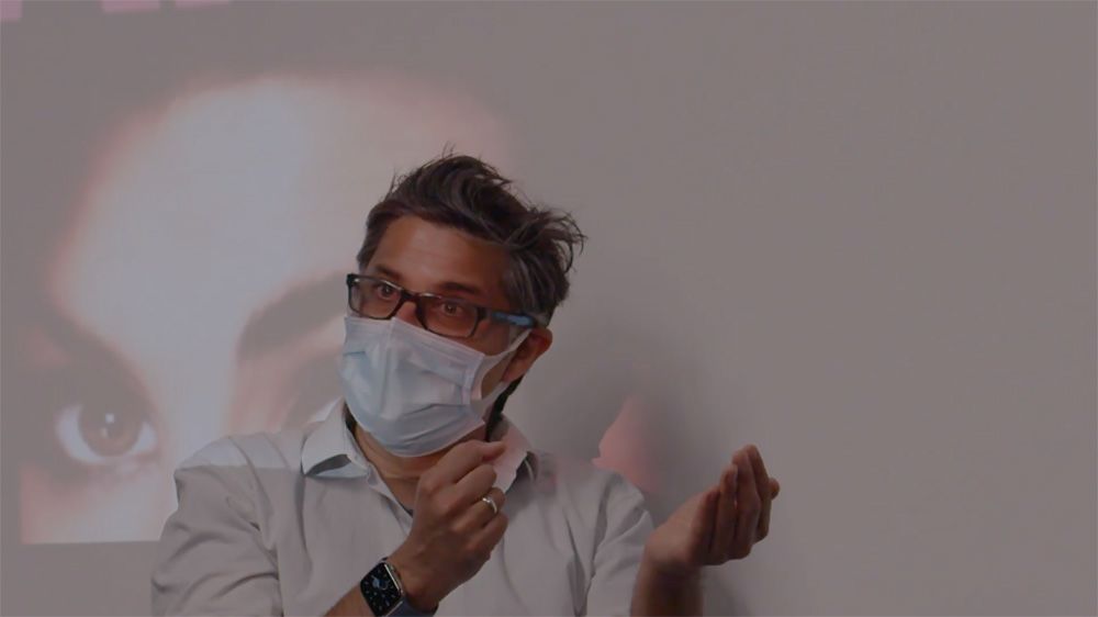 A director wearing a face mask sits in front of a projected screen.