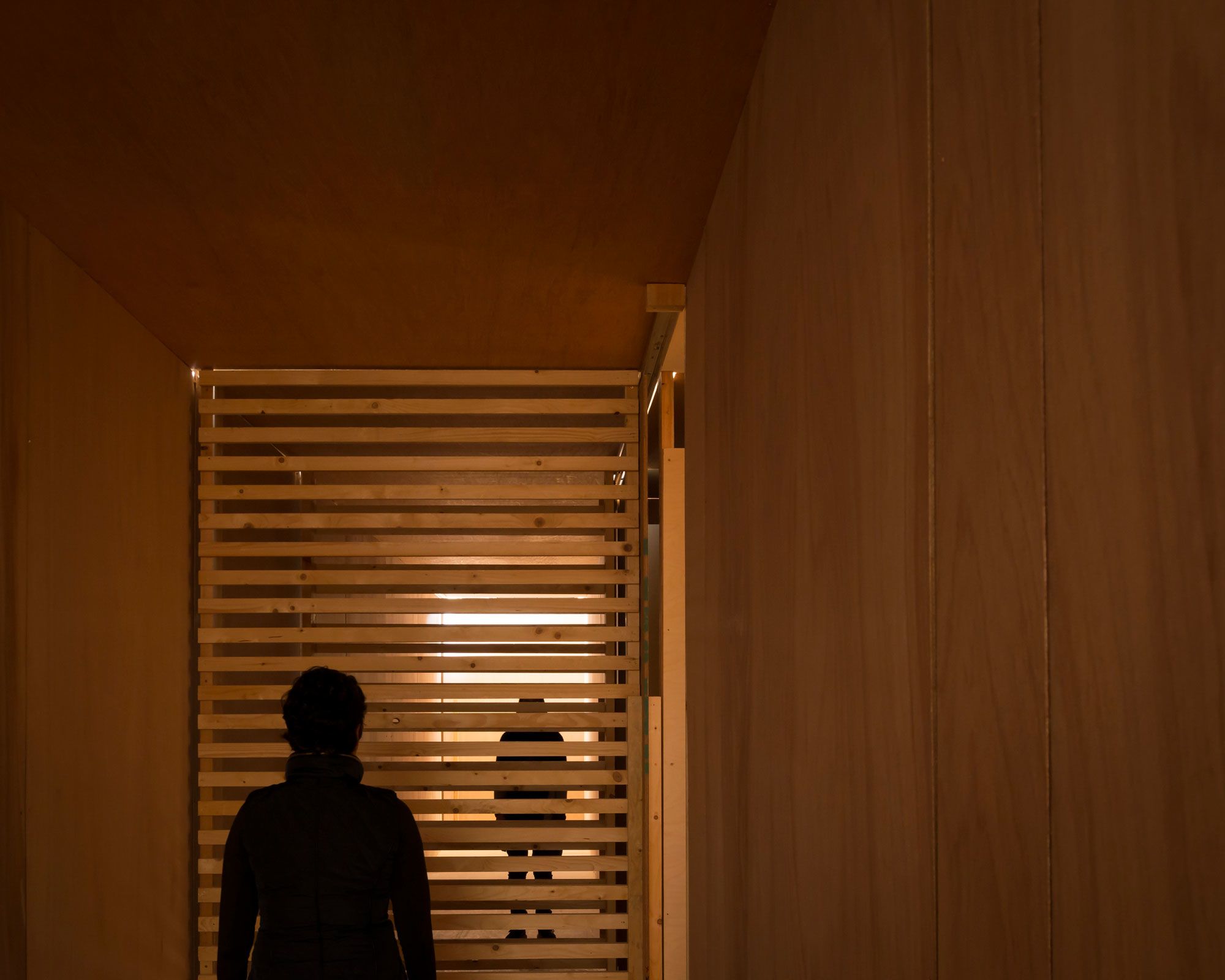 The silhouette of a man standing in a dimly lit wooden room with a slatted window/opening, another person is visible through the slats in an adjoining room.