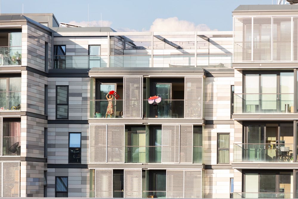 A shot of an apartment block with a figure in a dress on one of the balconies holding upright a red chair