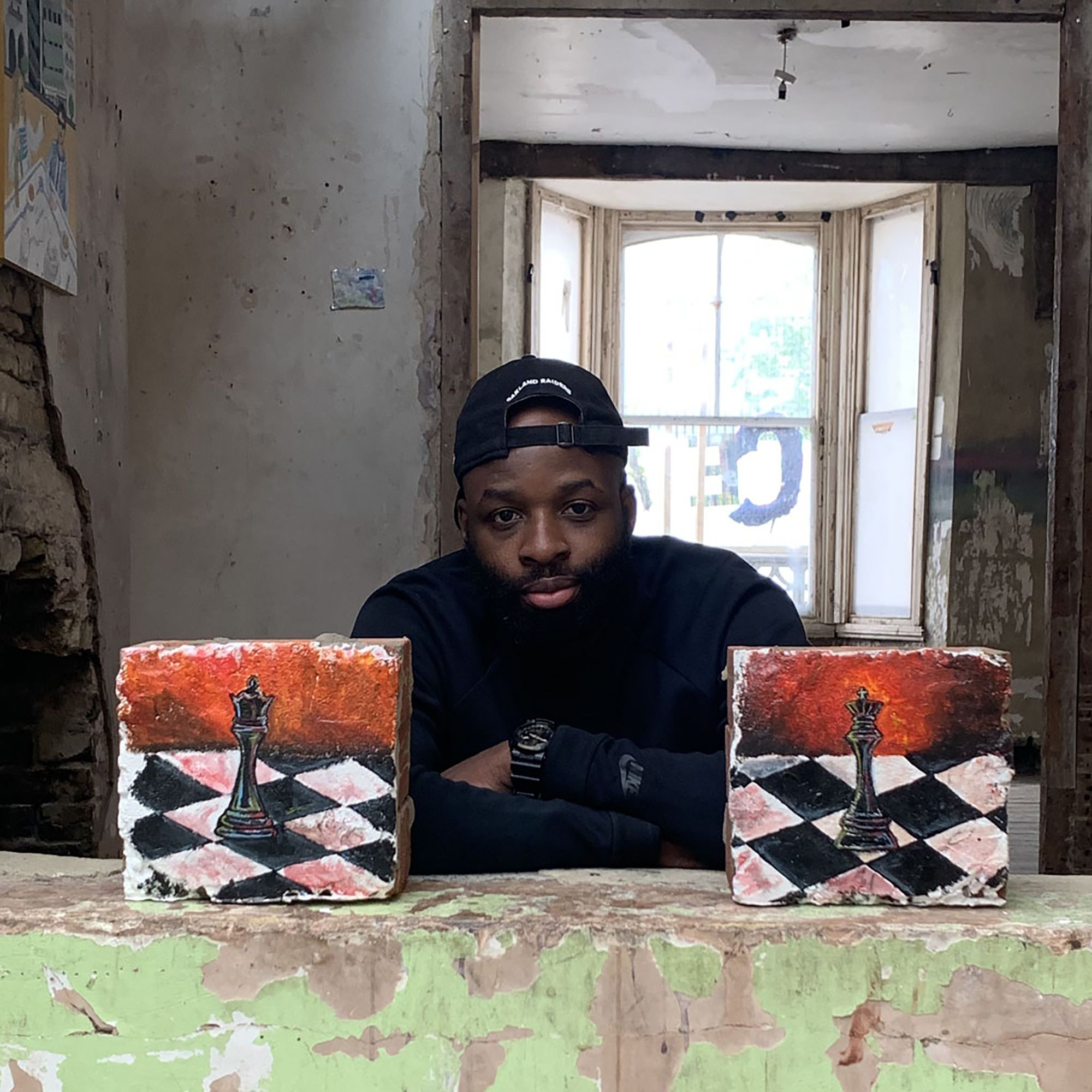 oseph, a black male, wearing all black clothing, wearing a backwards baseball cap is looking towards the camera, crossing his arms. He is positioned in between two of his artworks, paintings of chess pieces.  