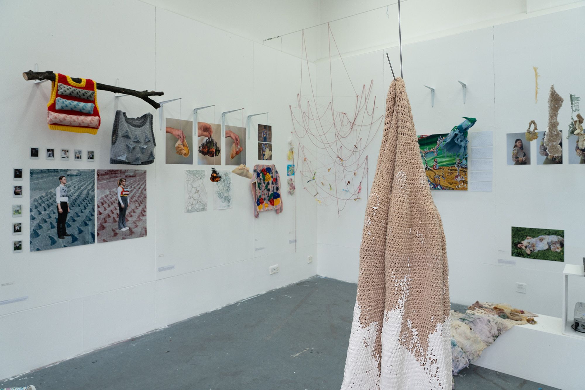A room shot of work from the Textile Craft specialism with a mix of work hung against a plain white wall. A large brown and white knitted blanket hangs from the ceiling and is touching the grey floor.  