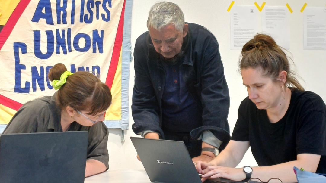 Image shows 3 people working round a table. 2 people are on laptops and 1 person is standing between them engaged in conversation.