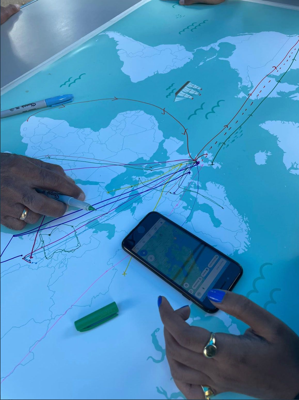 Two people's hands rest on a map of the world which has pen lines on it tracing routes from other countries to the UK. There is also a smart phone on the table.