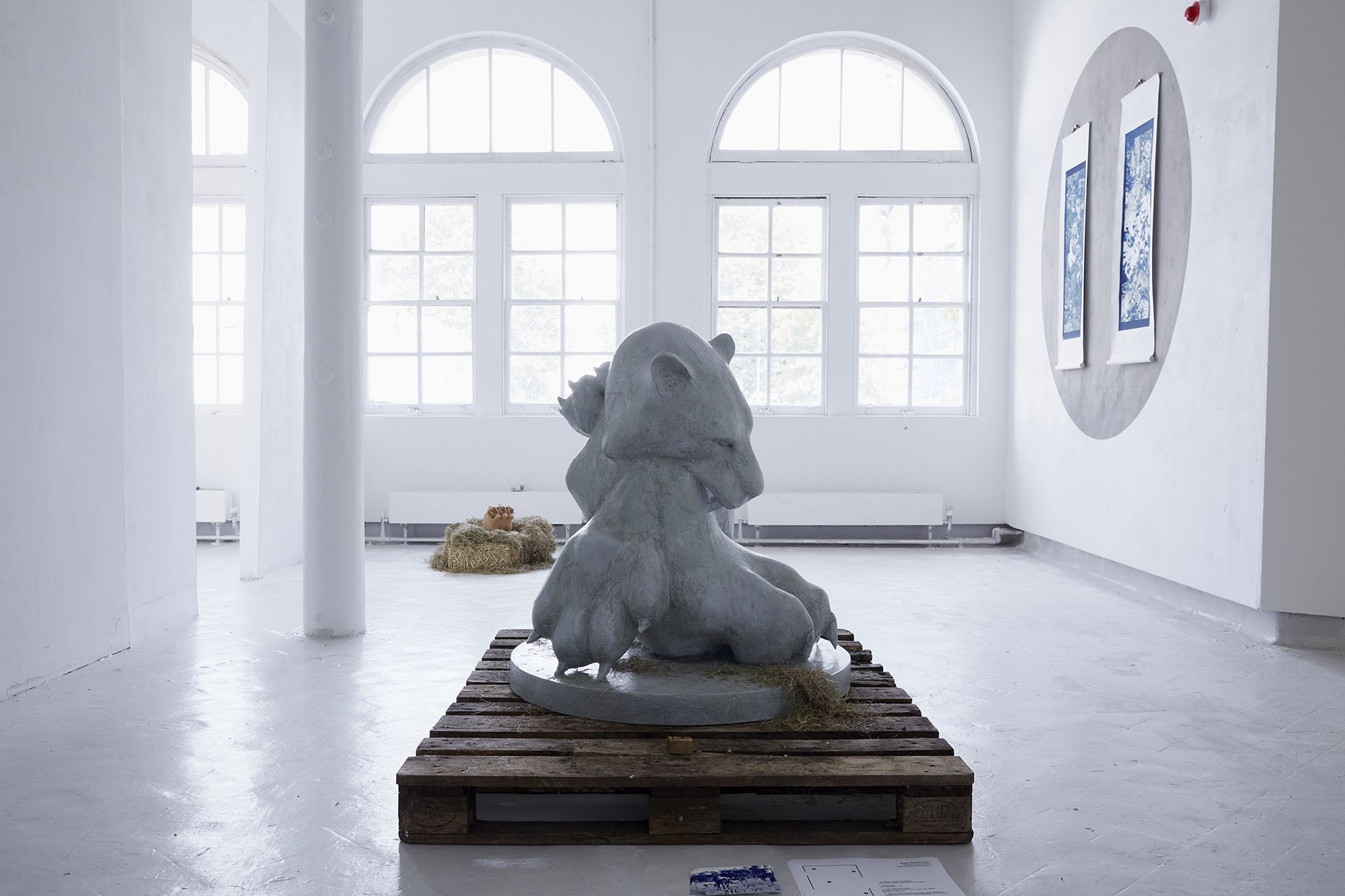 A sculpture of a large teddy like cat is sat on a brown wooden crate in a plain white studio. In the background there are two large windows and two pieces of art on the wall to the right. 