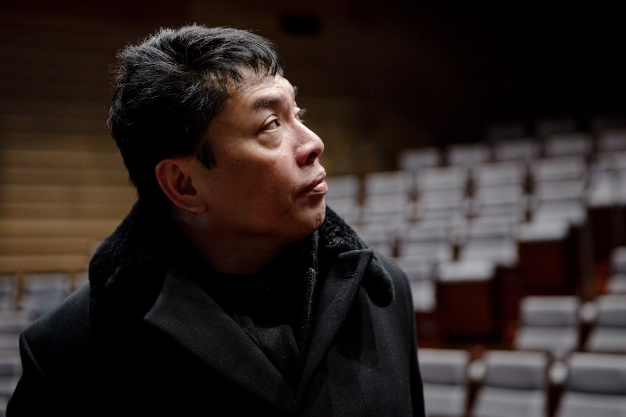 Image shows a man with black hair looking to the side against the backdrop of a theatre. He is wearing a black jacket and black scarf.