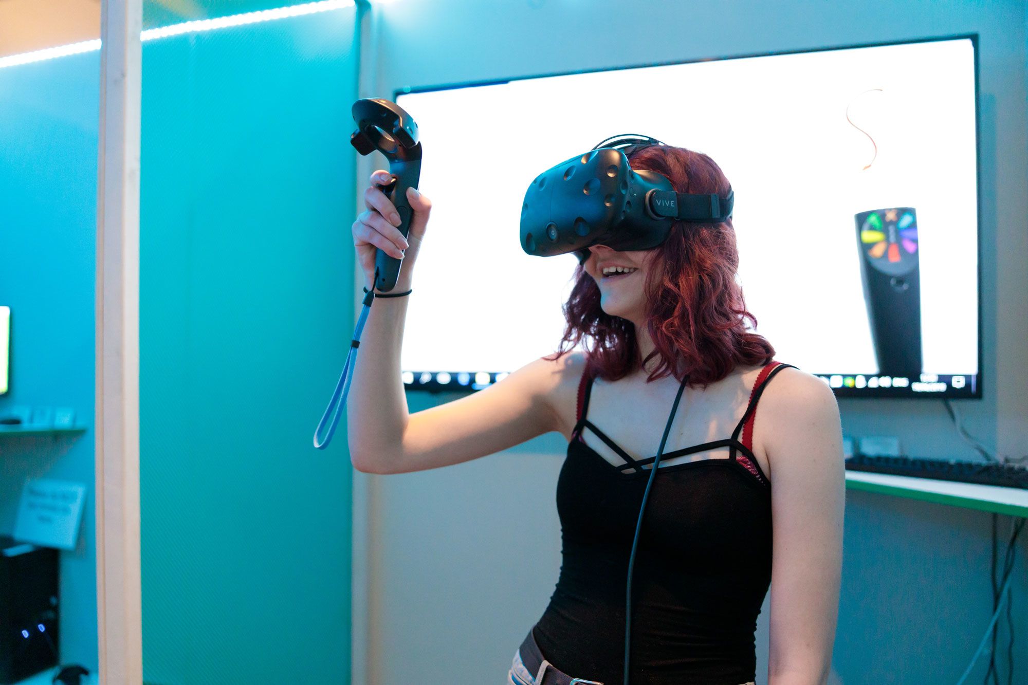 A girl in a black vest stop stands against a blue background wearing a VR headset and holding the controller in her hand, looking upwards.