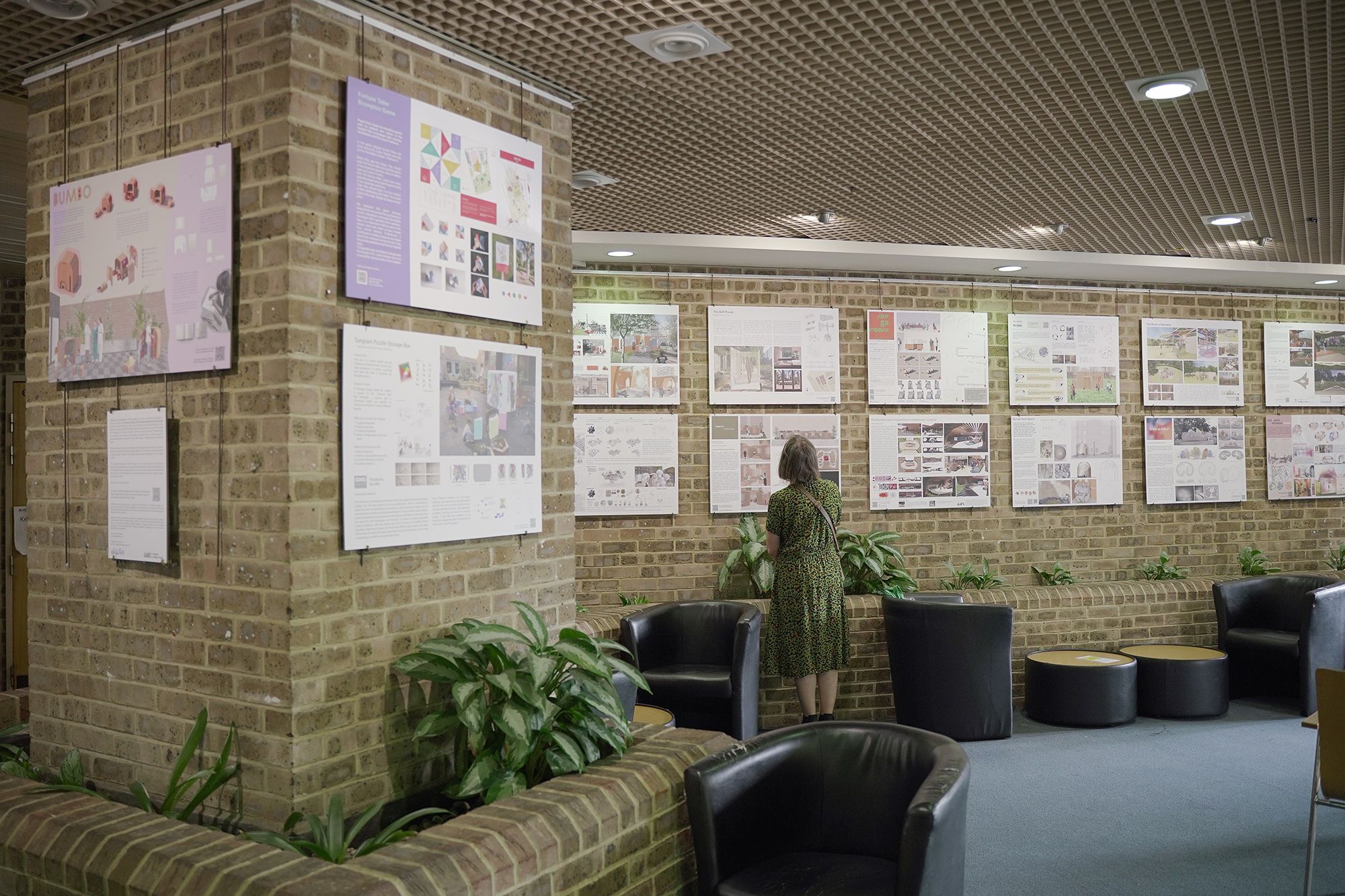  Photograph shows the final exhibition which took place at the Royal Brompton Hospital in the café space. The image shows a number of boards each with a design proposal for the hospital, there is a woman in a green dress looking at the boards. Around her are tables and chairs as well as some plants.  