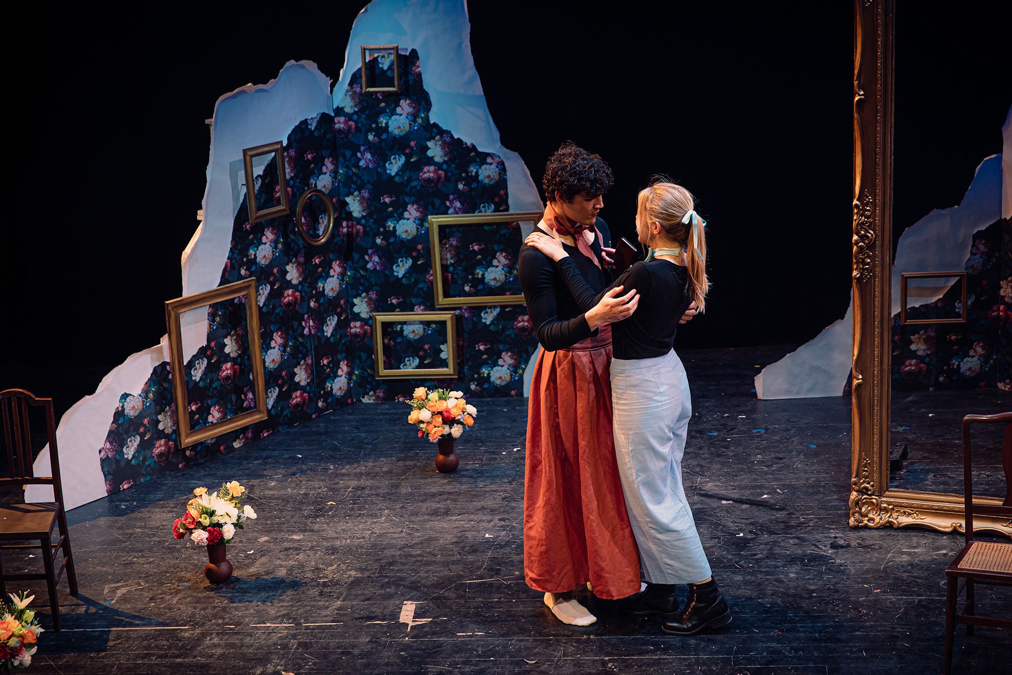 A stage with 2 people embracing. The set behind them contains a small triangle shaped wall with floral wallpaper and empty hanging gold picture frames. On the floor are 3 vases of flowers. 