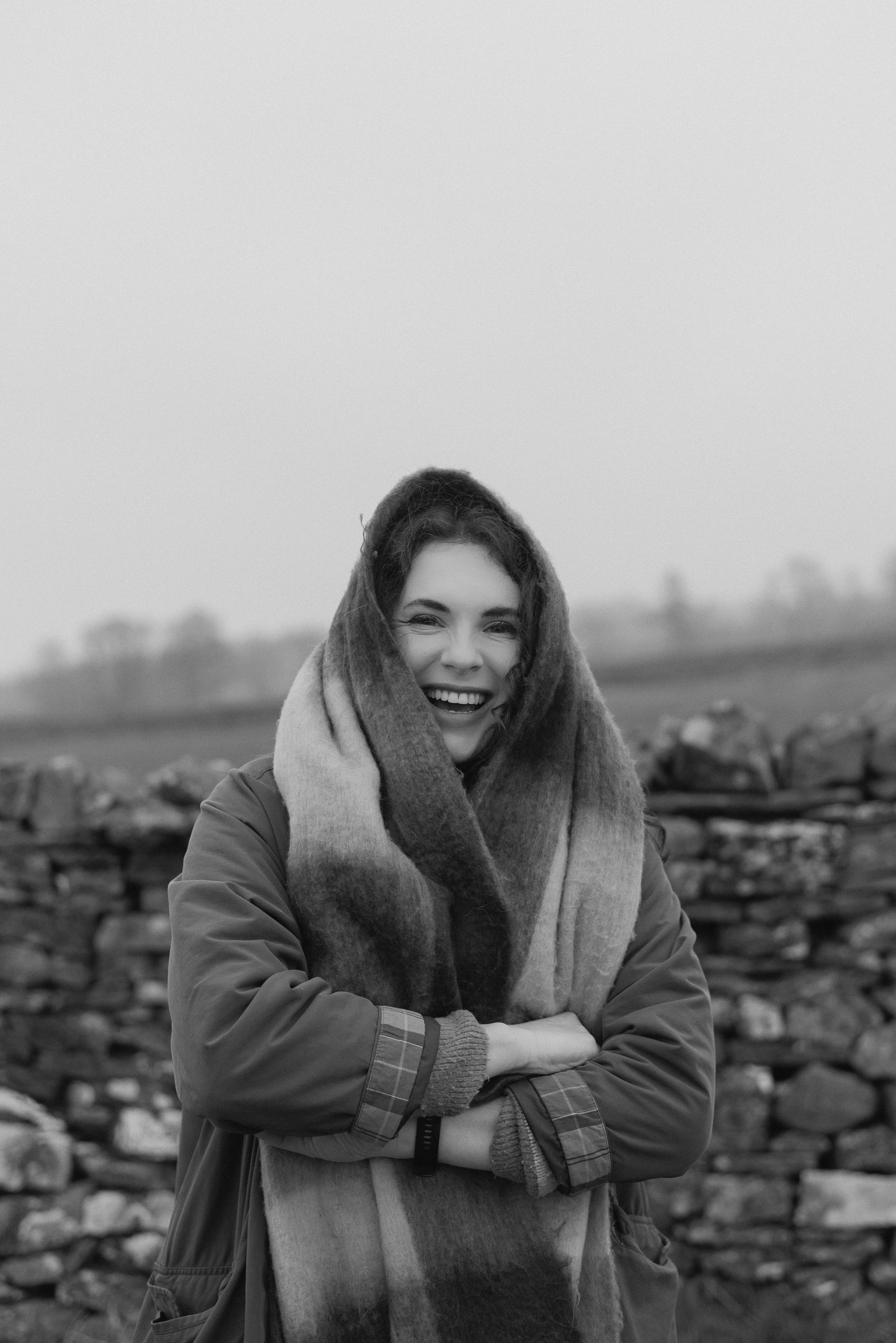 Black and white photo of Joanne smiling. She's got a bit scarf wrapped around her head and shoulders. Behind her is a dry stone wall