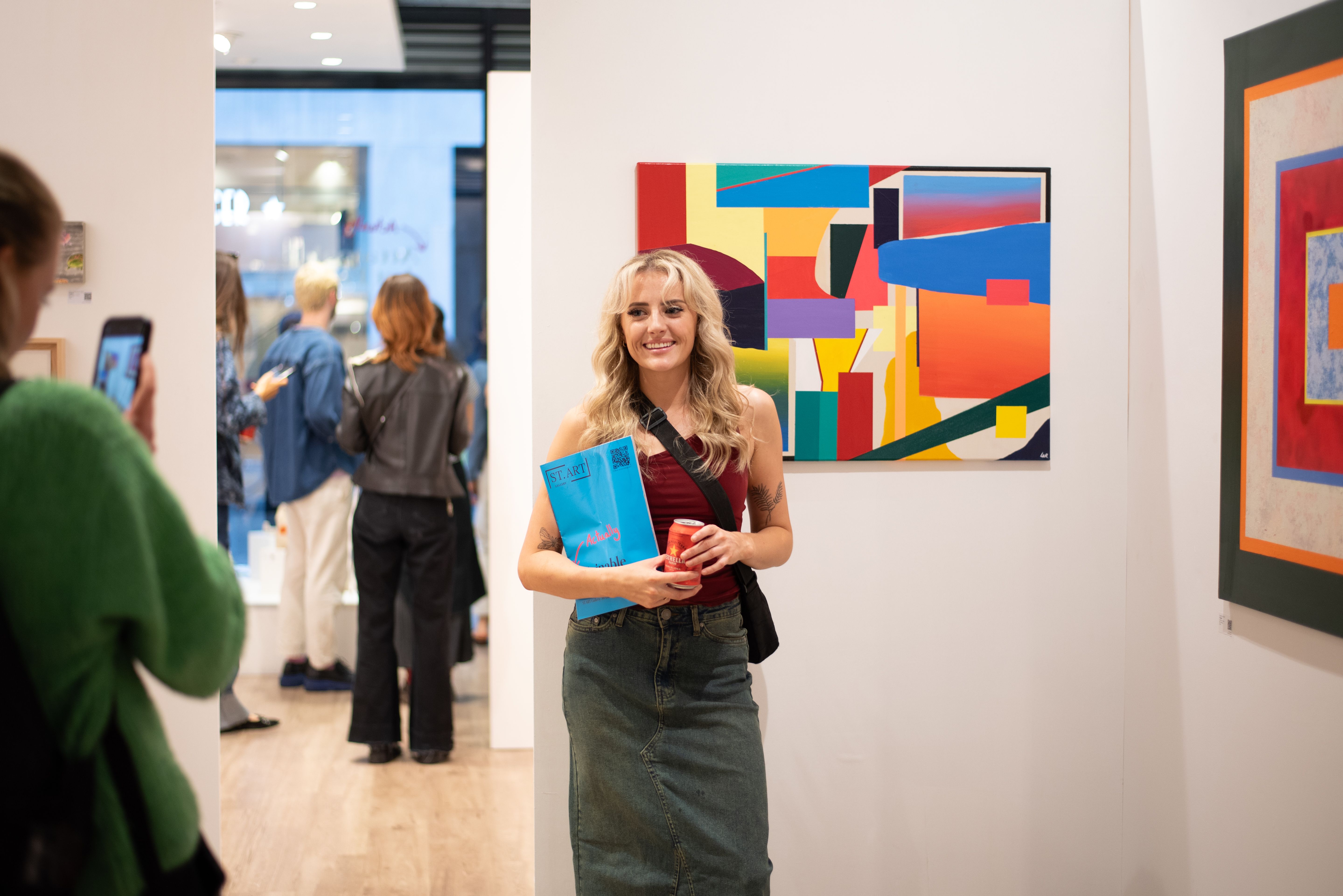 A woman, holding a ST.ART Gallery exhibition catalogue and a can. She's walking towards the camera, smiling. Behind her is a large abstract painting.