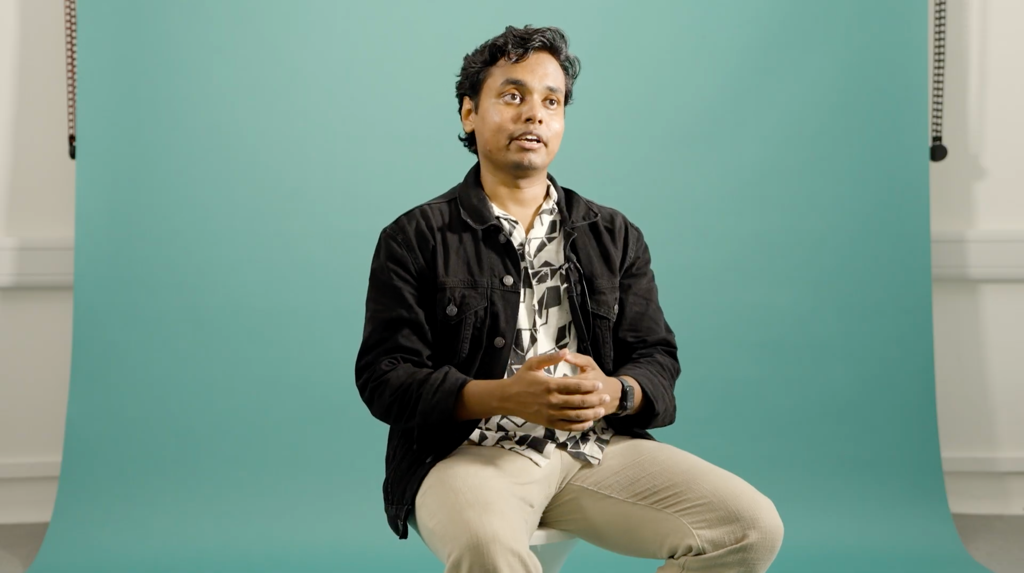 A young Indian male talks about his work, sitting on a stool in a photography studio, with a teal blue backdrop.