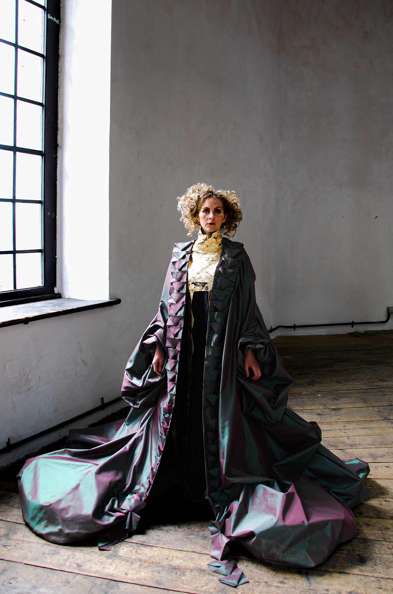  a woman with blonde curly hair, standing, modelling the costume, looking at the camera. Wearing a large purple/green coat which falls to the floor 