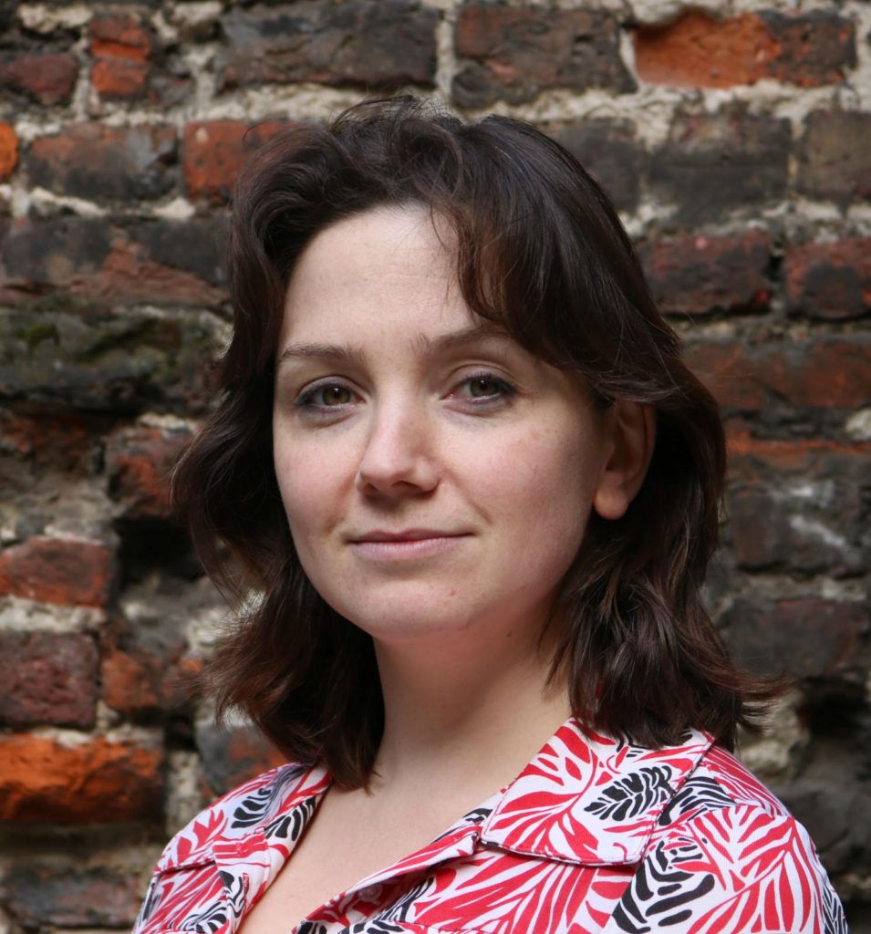 Headshot of Polly Mackenzie who is stood in front of a red brick wall