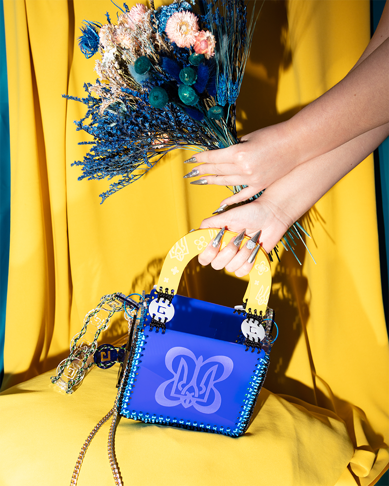 A blue handbag with a print and yellow handle, against a yellow garment. 