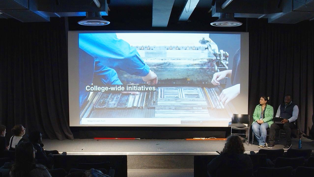 LCC Head of College Kene Igweonu and LCC Officer for Arts SU Sophia Nasif sit on stage at LCC in front of a projection with the text 'College-wide initiatives' and an image of hands over a typesetting machine