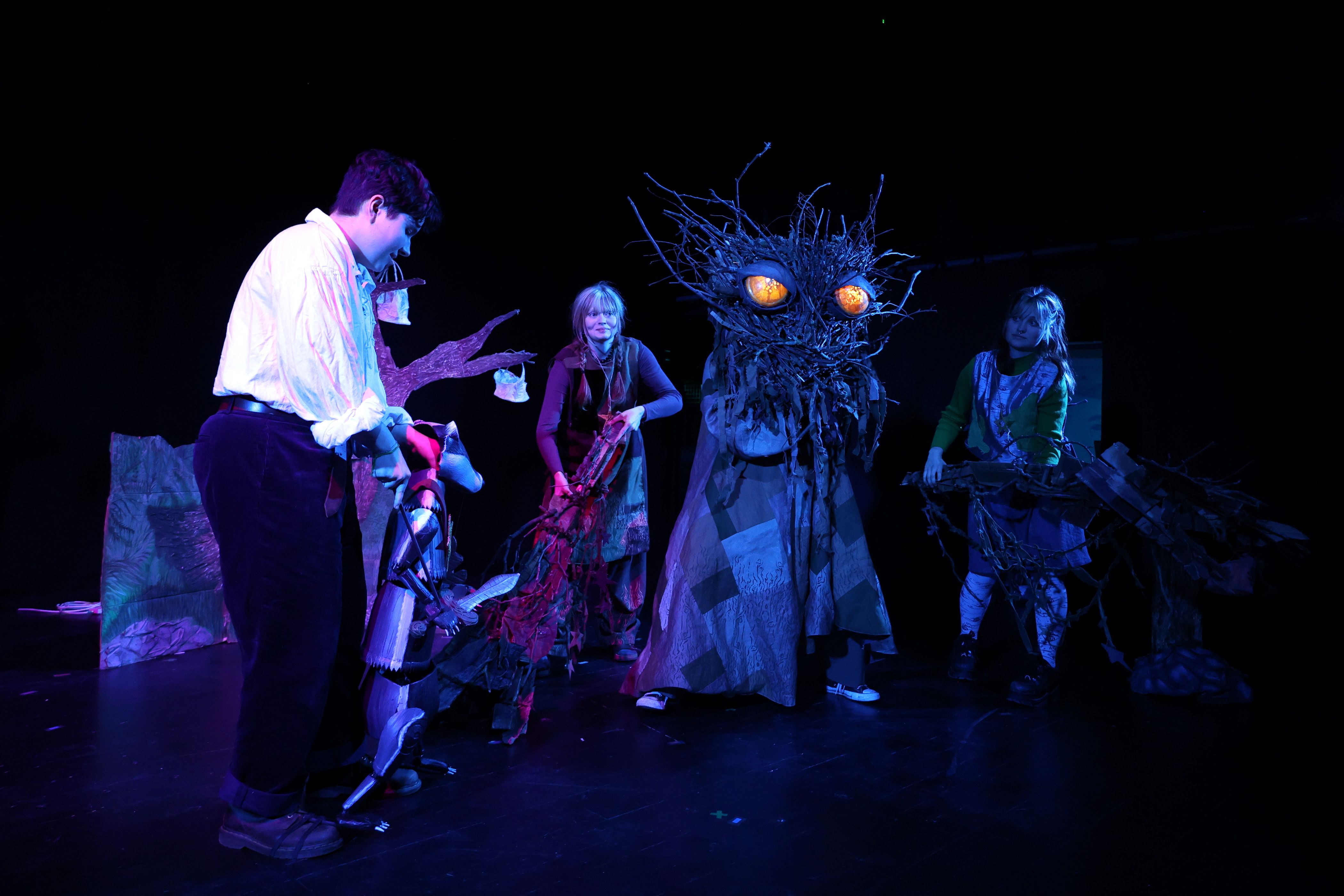 Images show 3 performers on stage with a 4th actor wearing a large wooden headpiece with yellow glowing eyes.