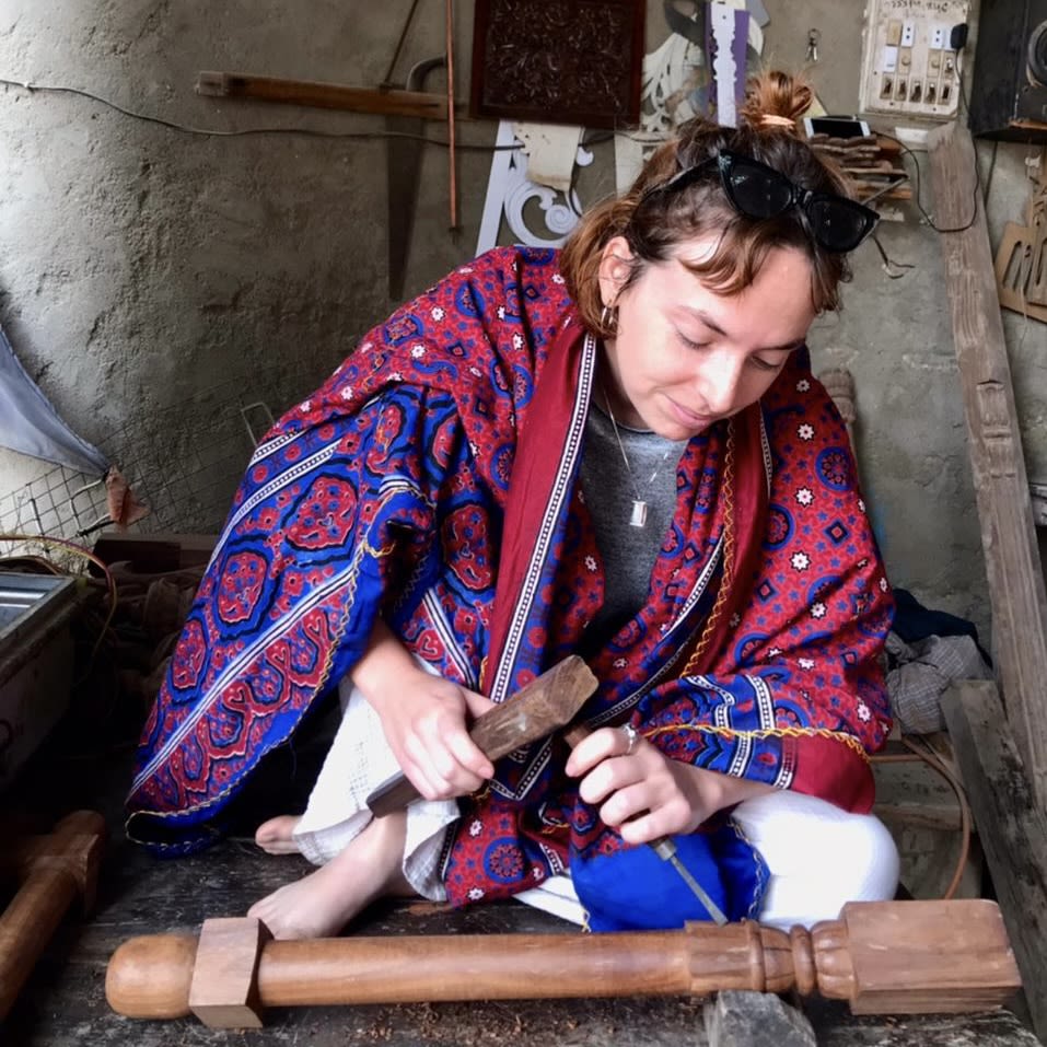 Nele Bergmans sits on the floor wearing a red and blue shawl carving a wooden leg for the stand of her piece using traditional hand crafting tools.  