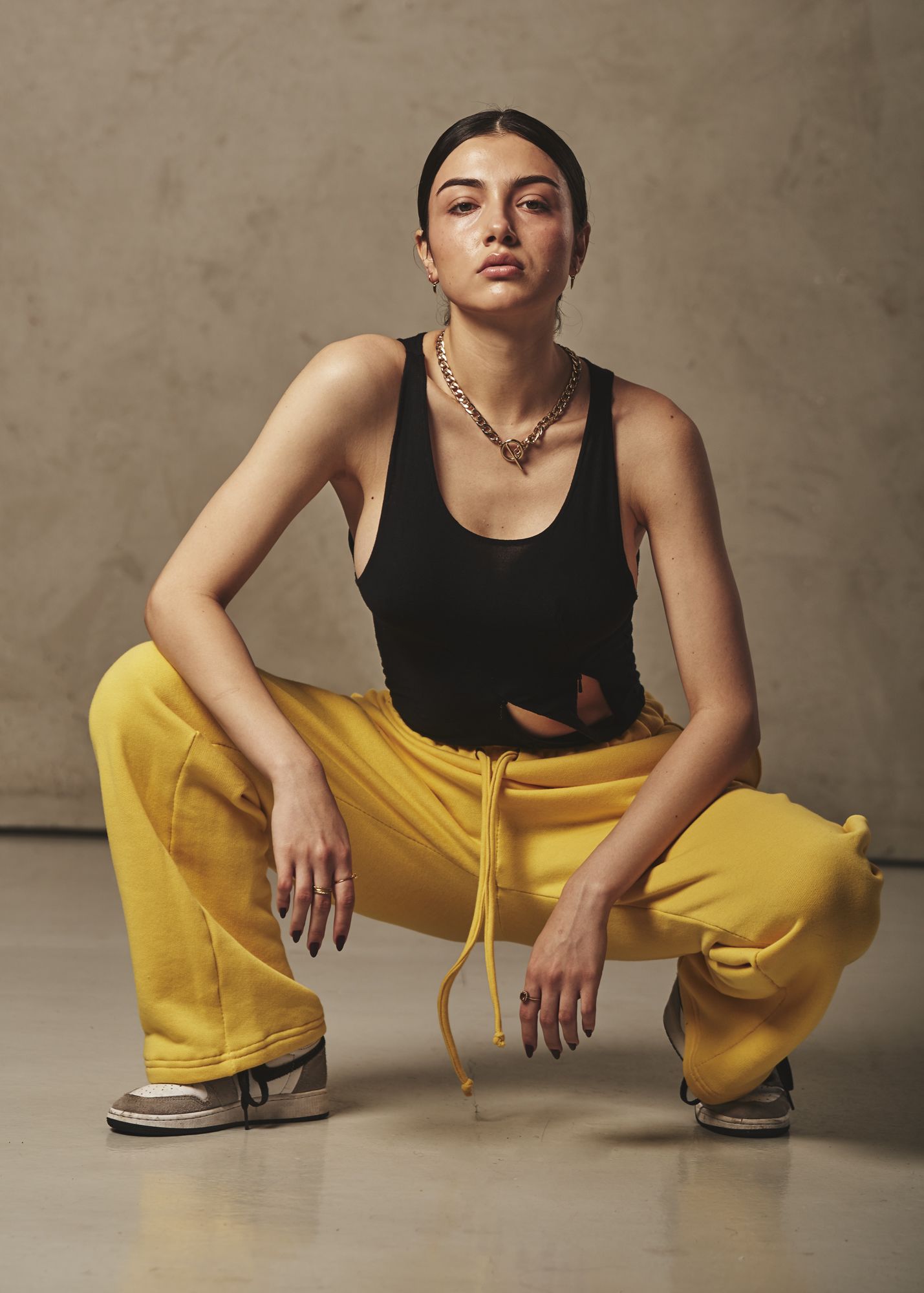 A woman modelling athletic attire, comprising yellow sweatpants, a black tank top with cut outs, and a gold chain neckalce. The model is crouched in a squatted position