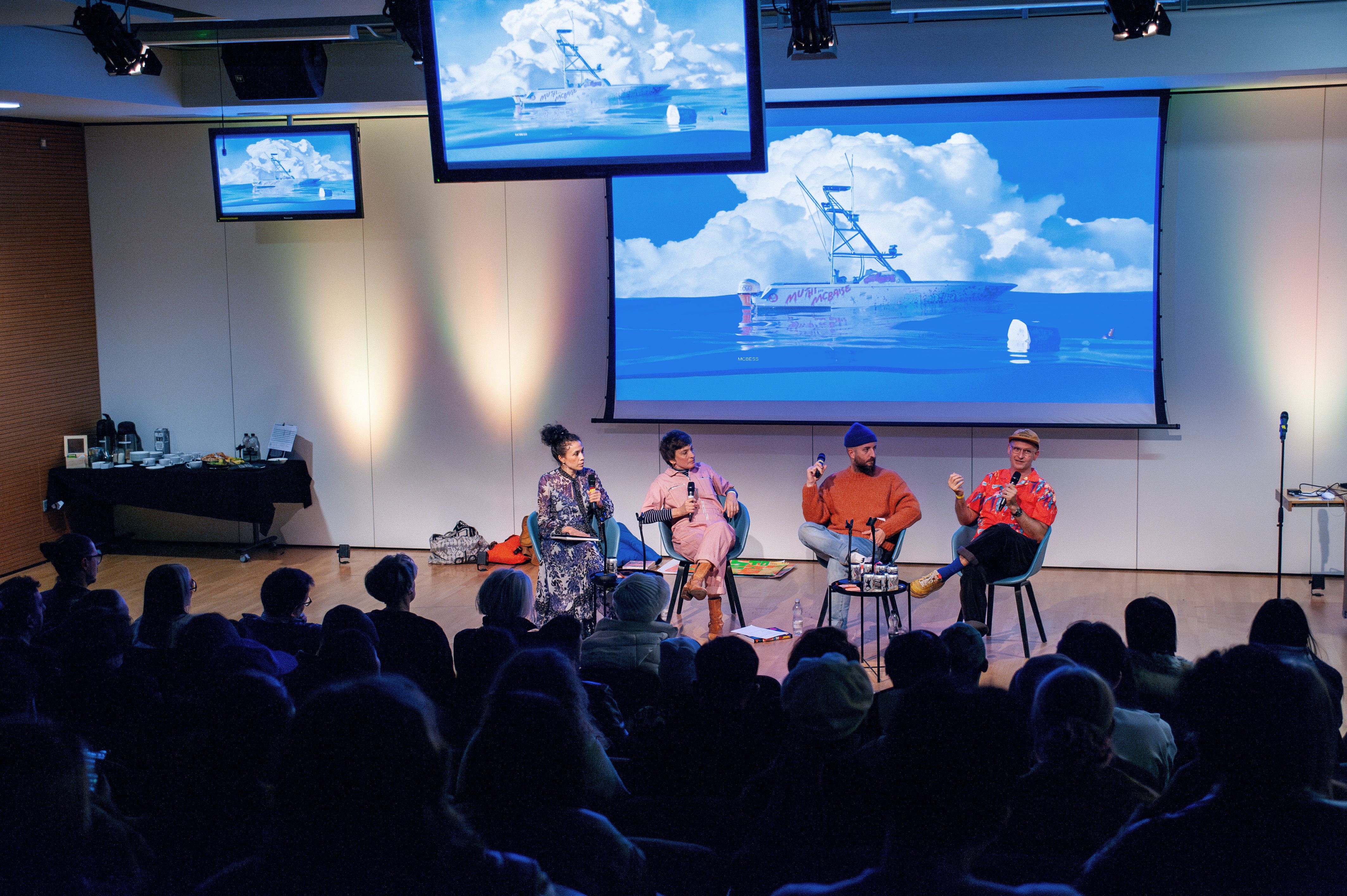 Wide shot from the back of the room, with the guests sitting facing the front of the room and the panellists at the front. There's screens above them.