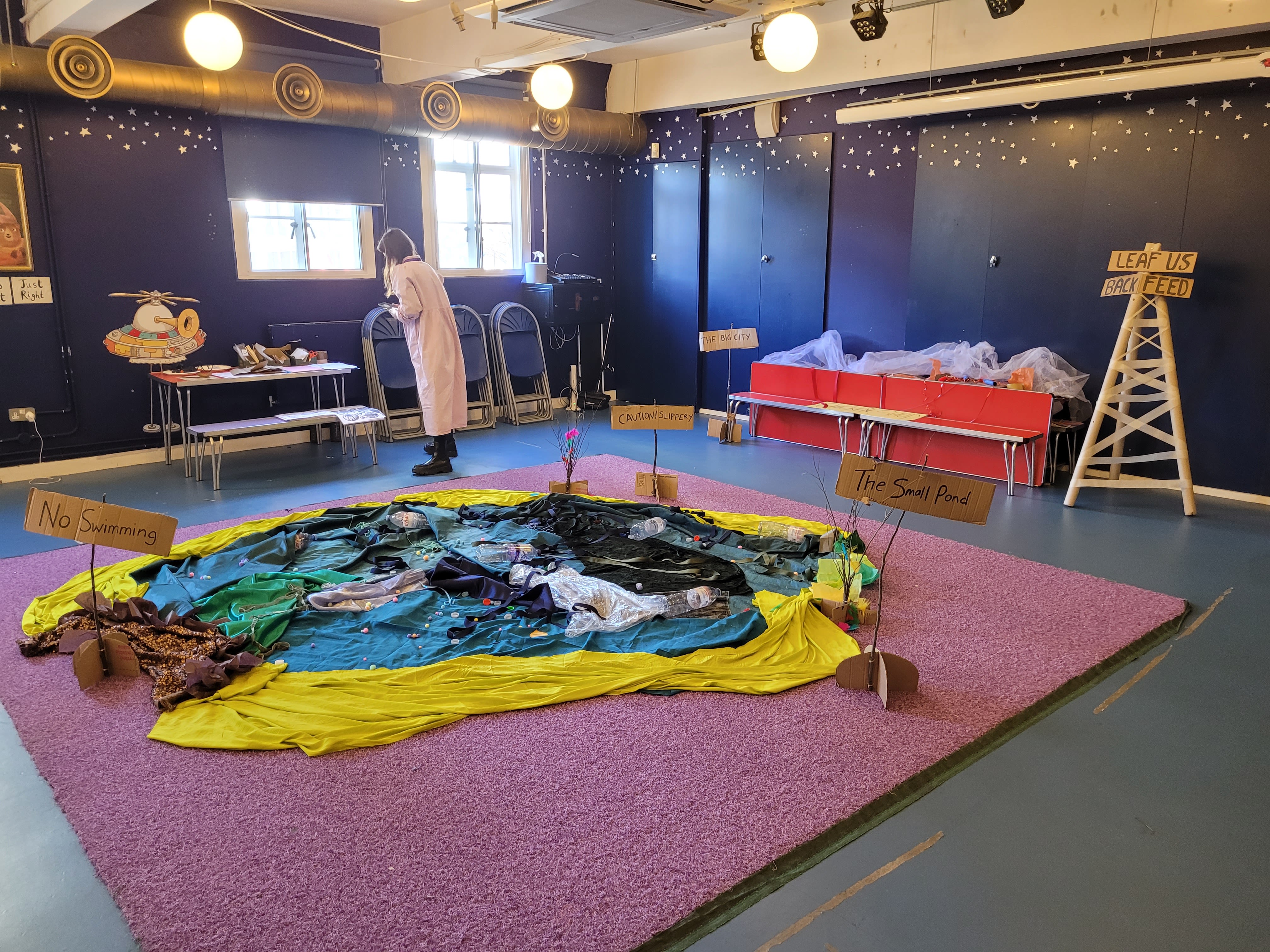 Large room with blue wallpaper and golden stars decorated on the walls. In centre of room, colourful materials displayed in river/bog like shape.