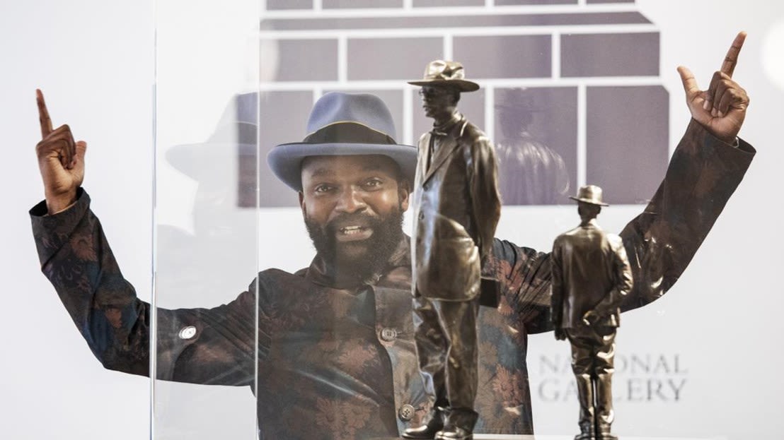 A Black man with his hands up with fingers pointing upwards in front of two bronze-coloured statues of men in hats. 