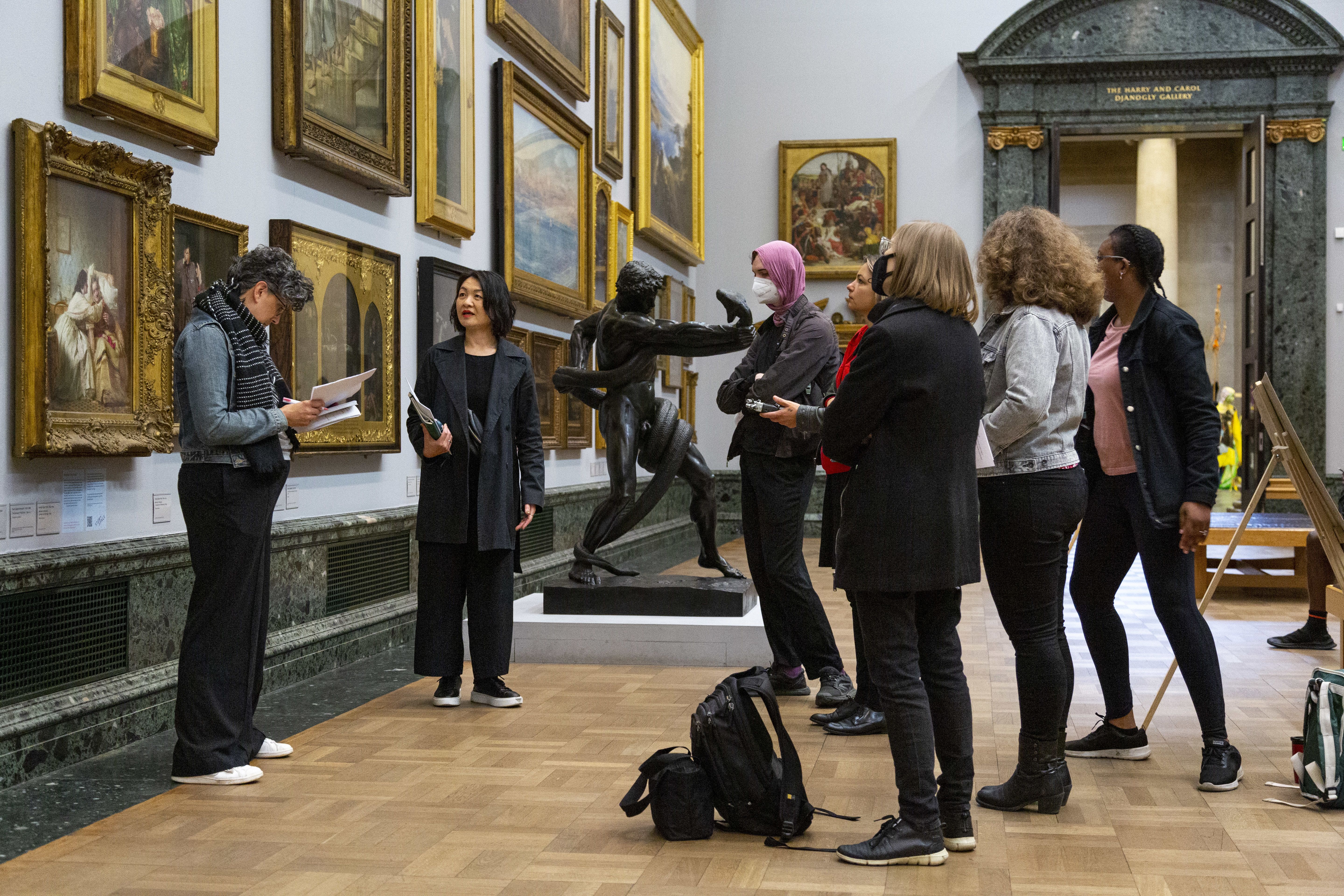 A group of people in a museum looking at ornately framed artworks on the wall. There is a parquet floor and a doorway in the background with pillars 