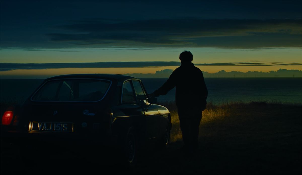 Still of a man standing beside a car at dawn.