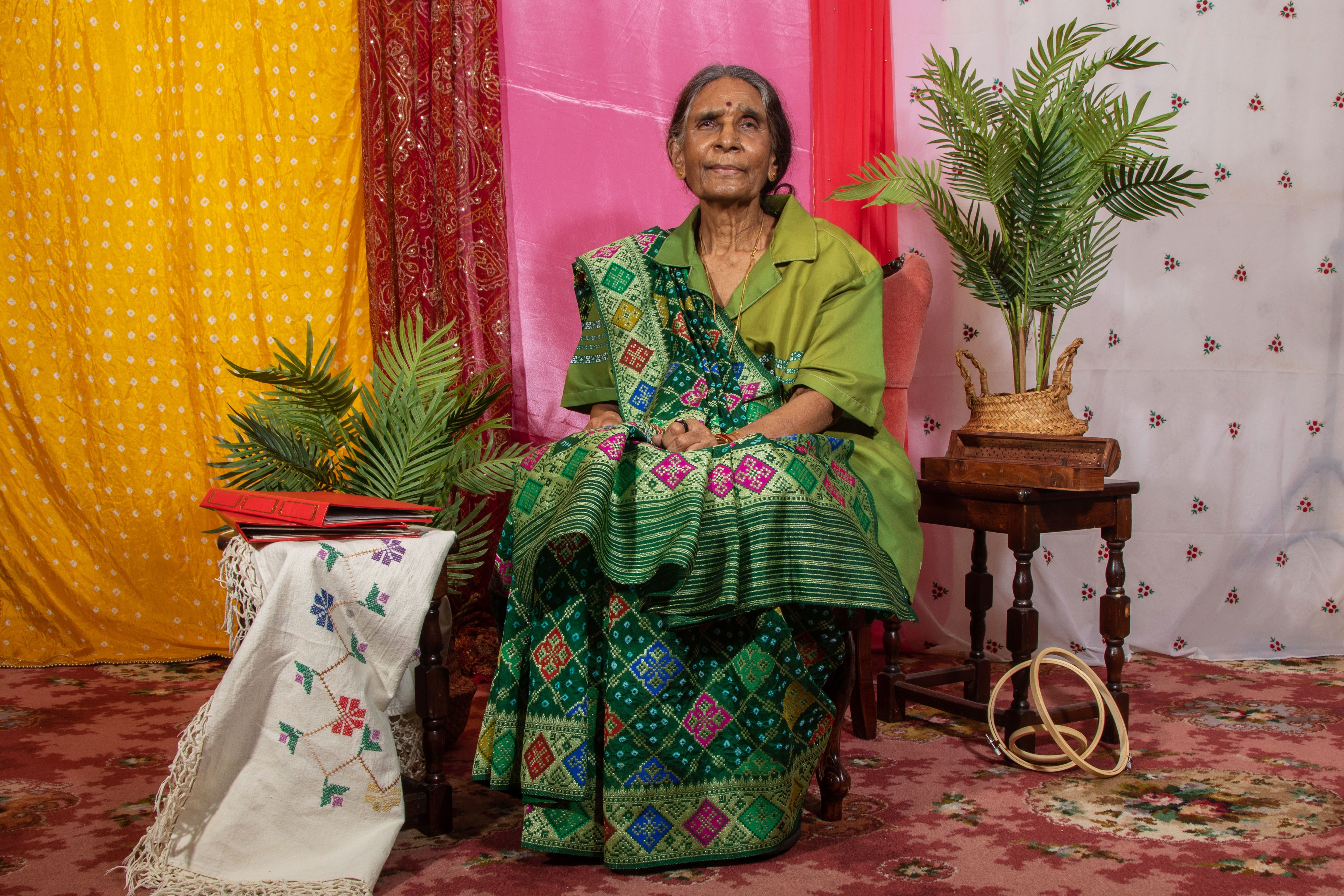 An elderly Sri Lankan woman wearing a sari in different tones of green. Different materials make-up the background and there's plants on either side