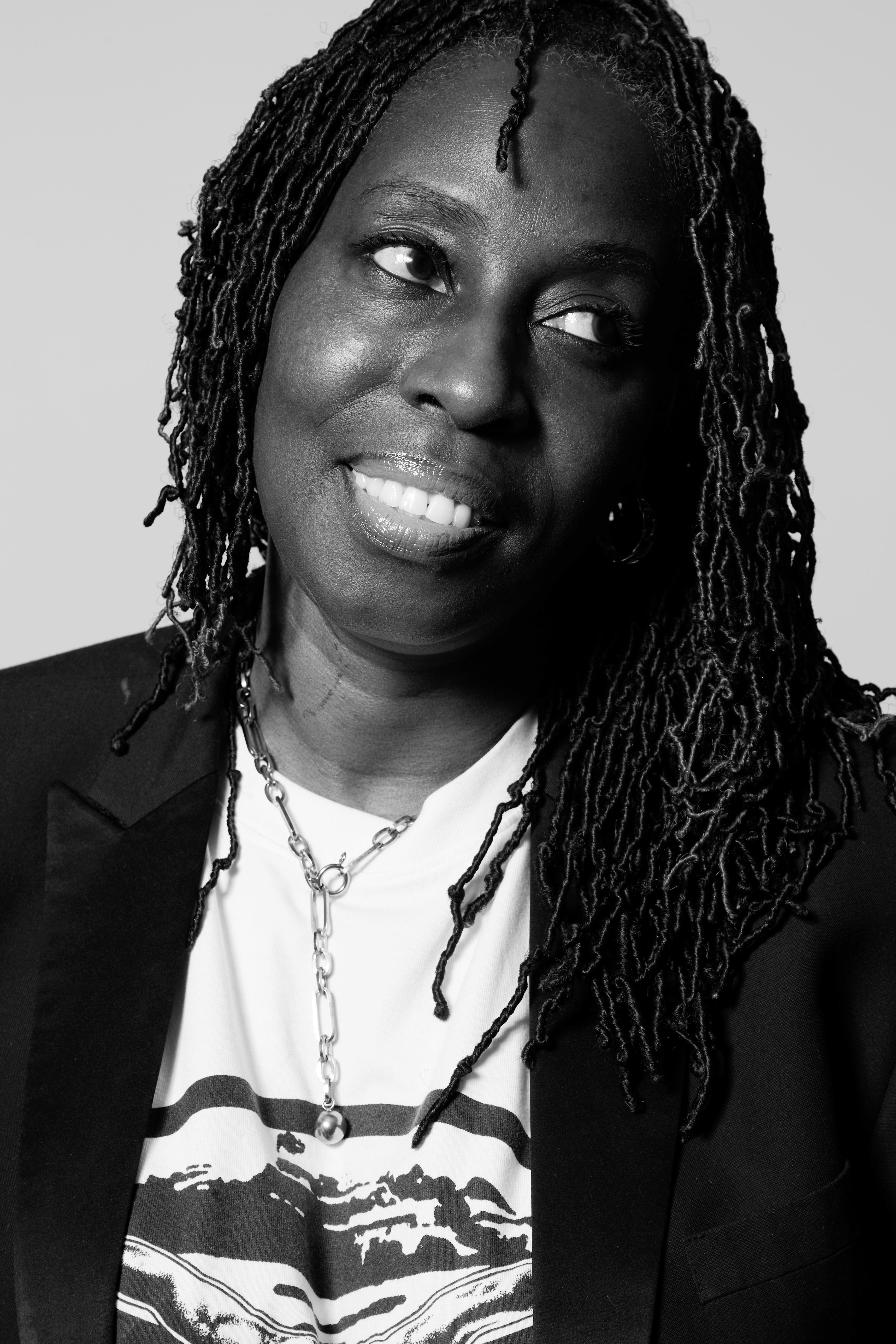 A person with long, textured hair wearing a dark blazer, a printed T‑shirt, and a chain necklace in a black‑and‑white portrait.