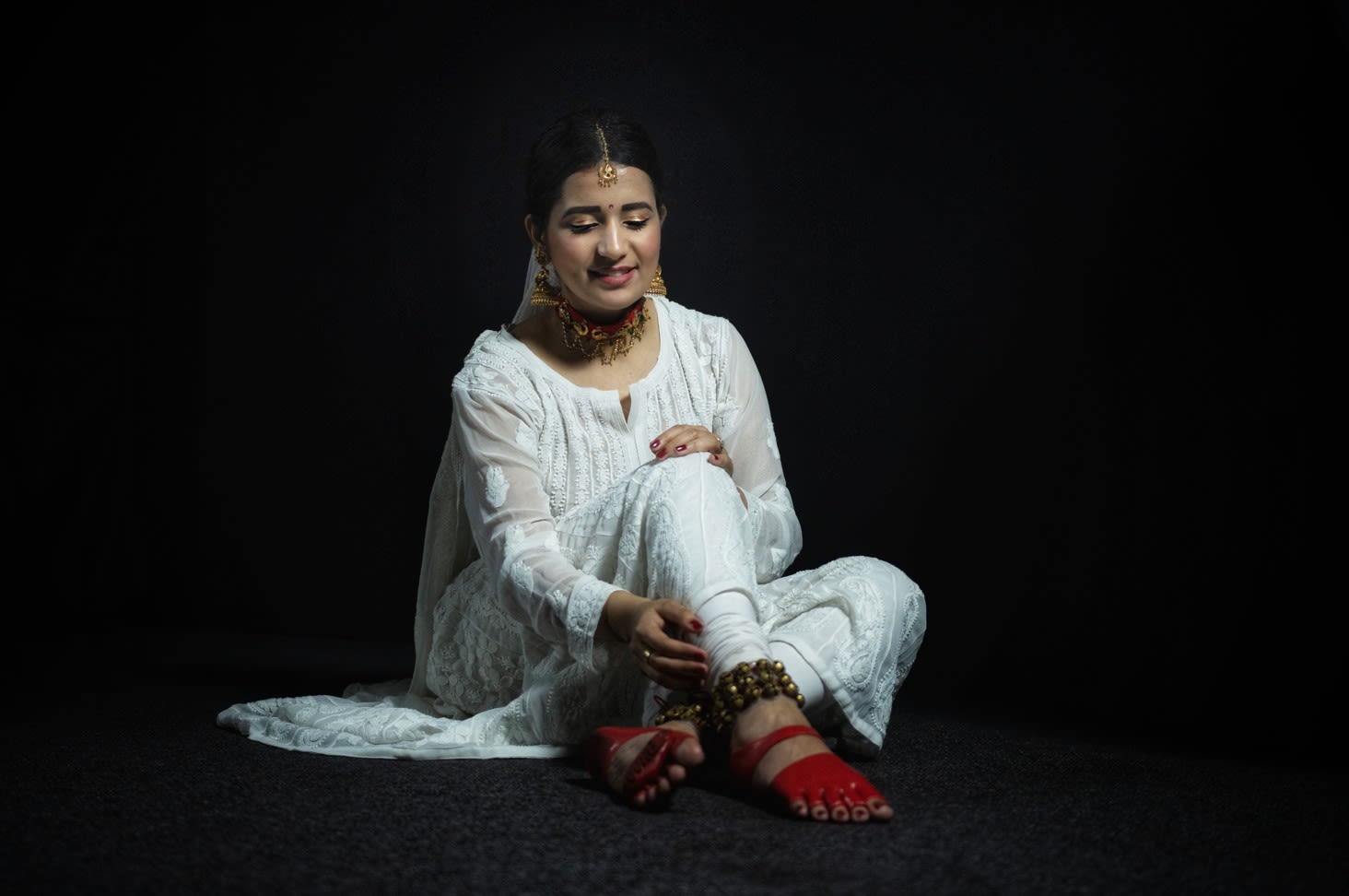Graduate student Aditi Sarna sitting down wearing her footwear design and white traditional dress with a black background