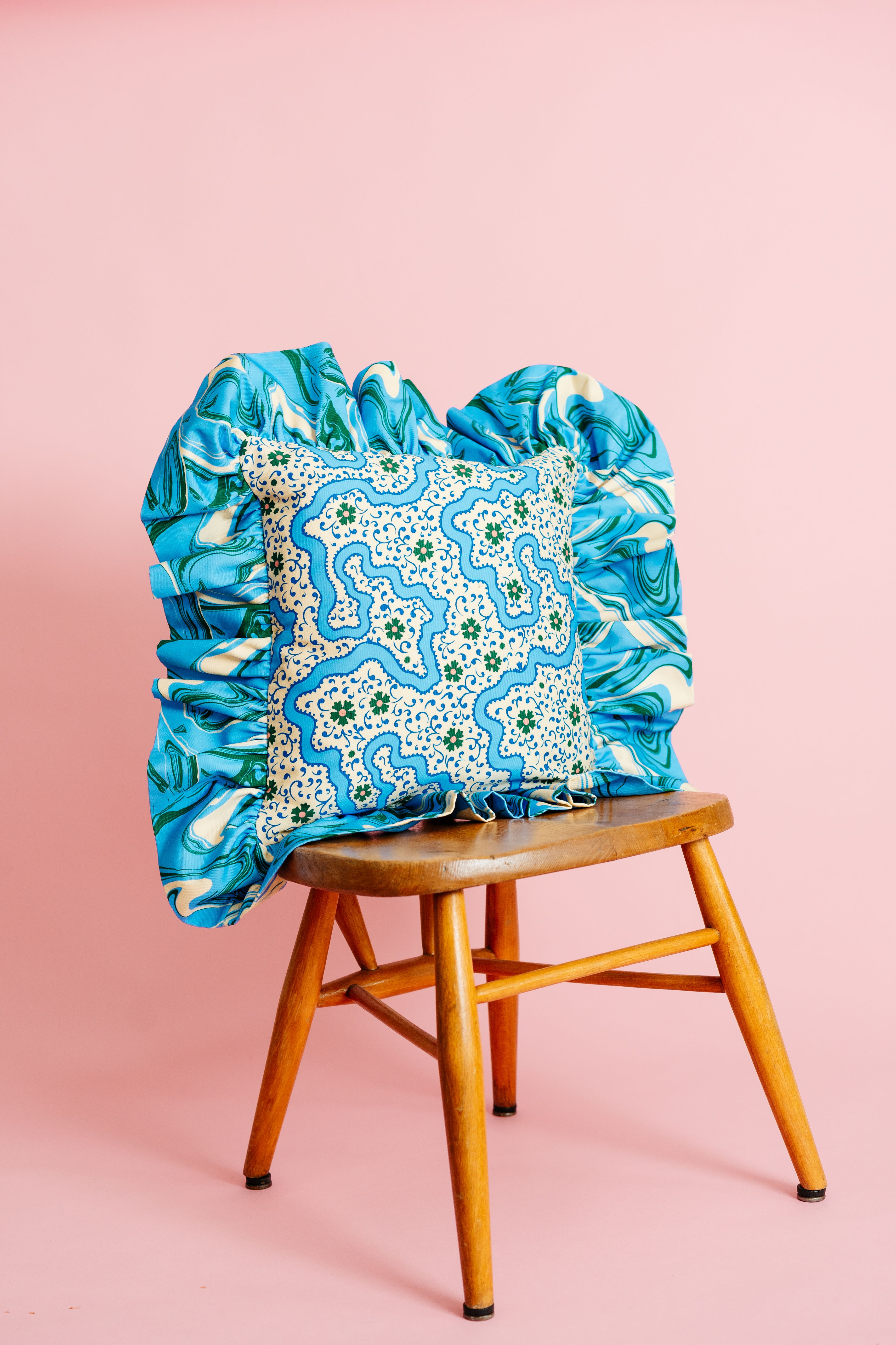 A blue and green patterned cushion resting on a wooden stool. The background of the photo is a pale pink