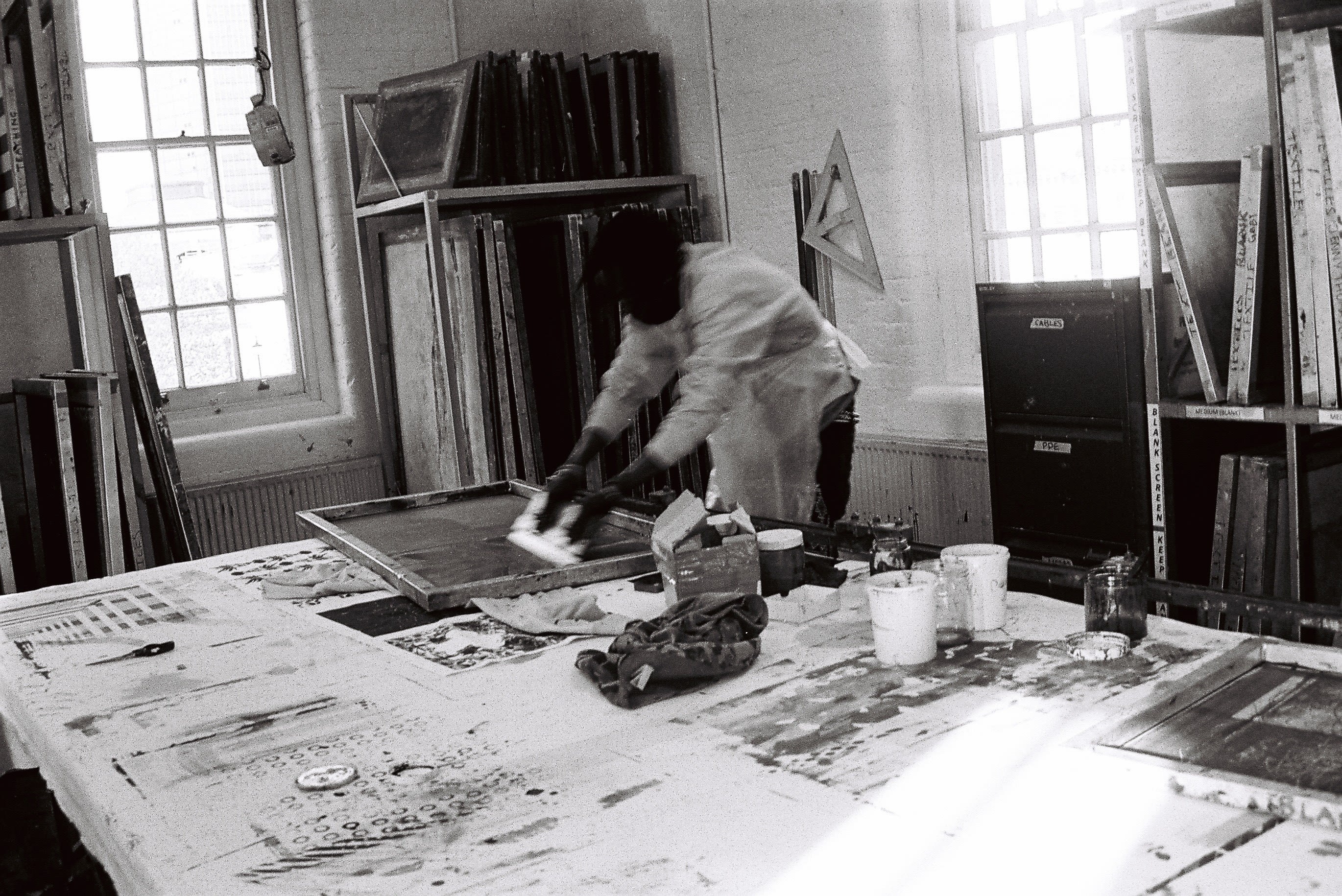 Image shows a person in a workshop Screenprinting work on a large table in front of them. In the background there are large windows 