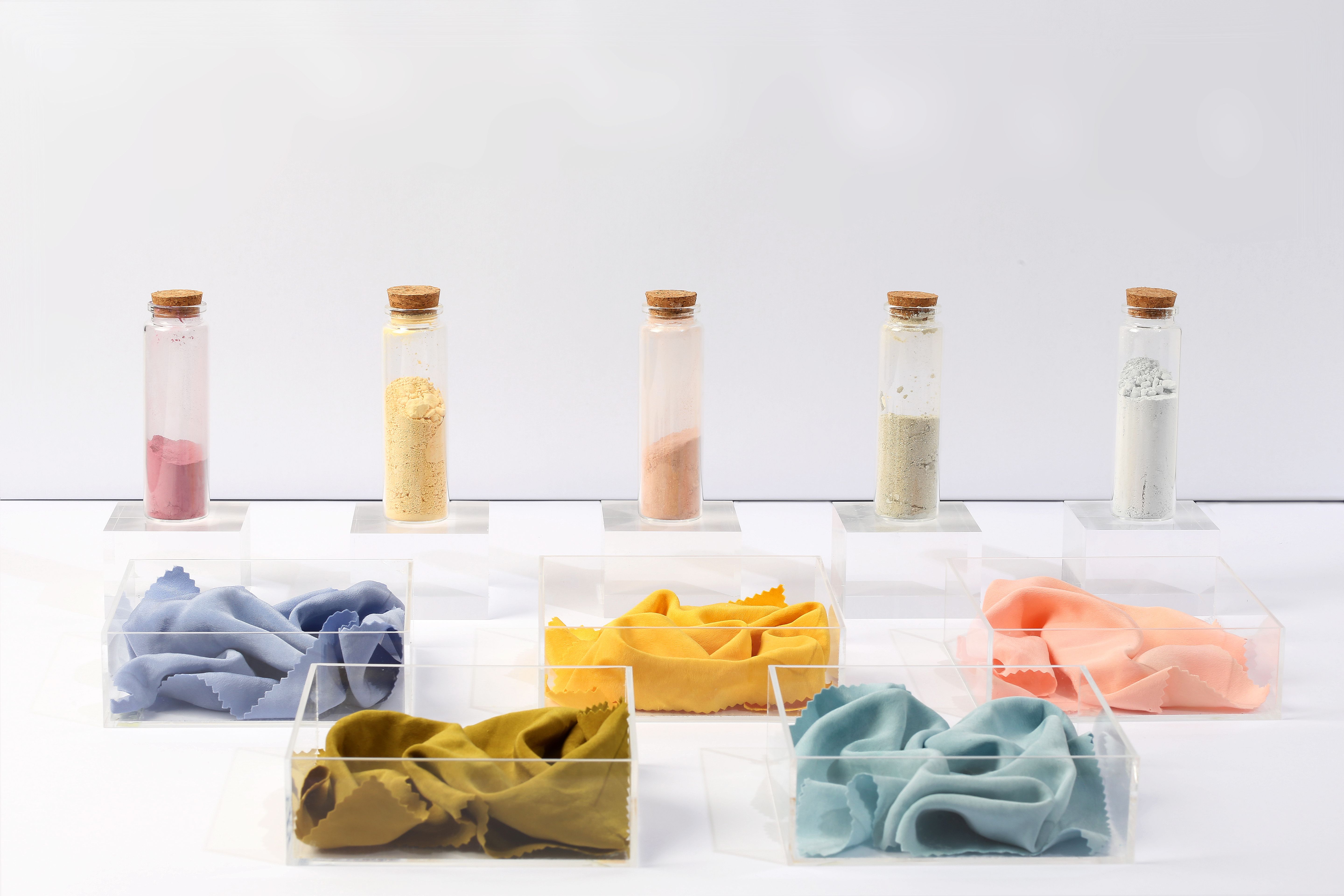 5 corked tubes with coloured dye powders within them, placed behind boxes with different coloured fabrics in, reflecting the dye pigments. 