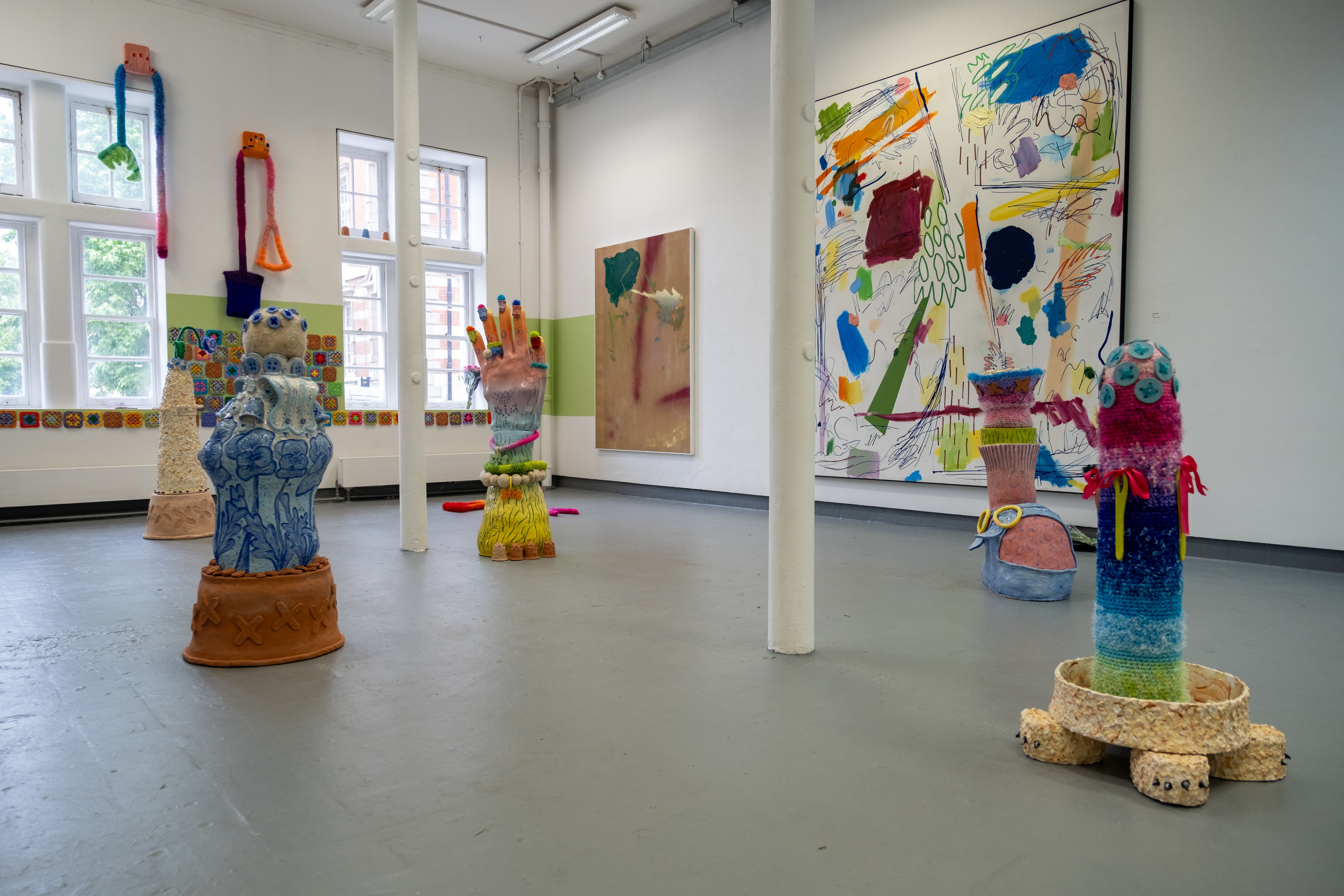 Image shows a series of colourful artworks sitting in a bright white studio room. On the walls, brightly coloured paintings can be seen. 