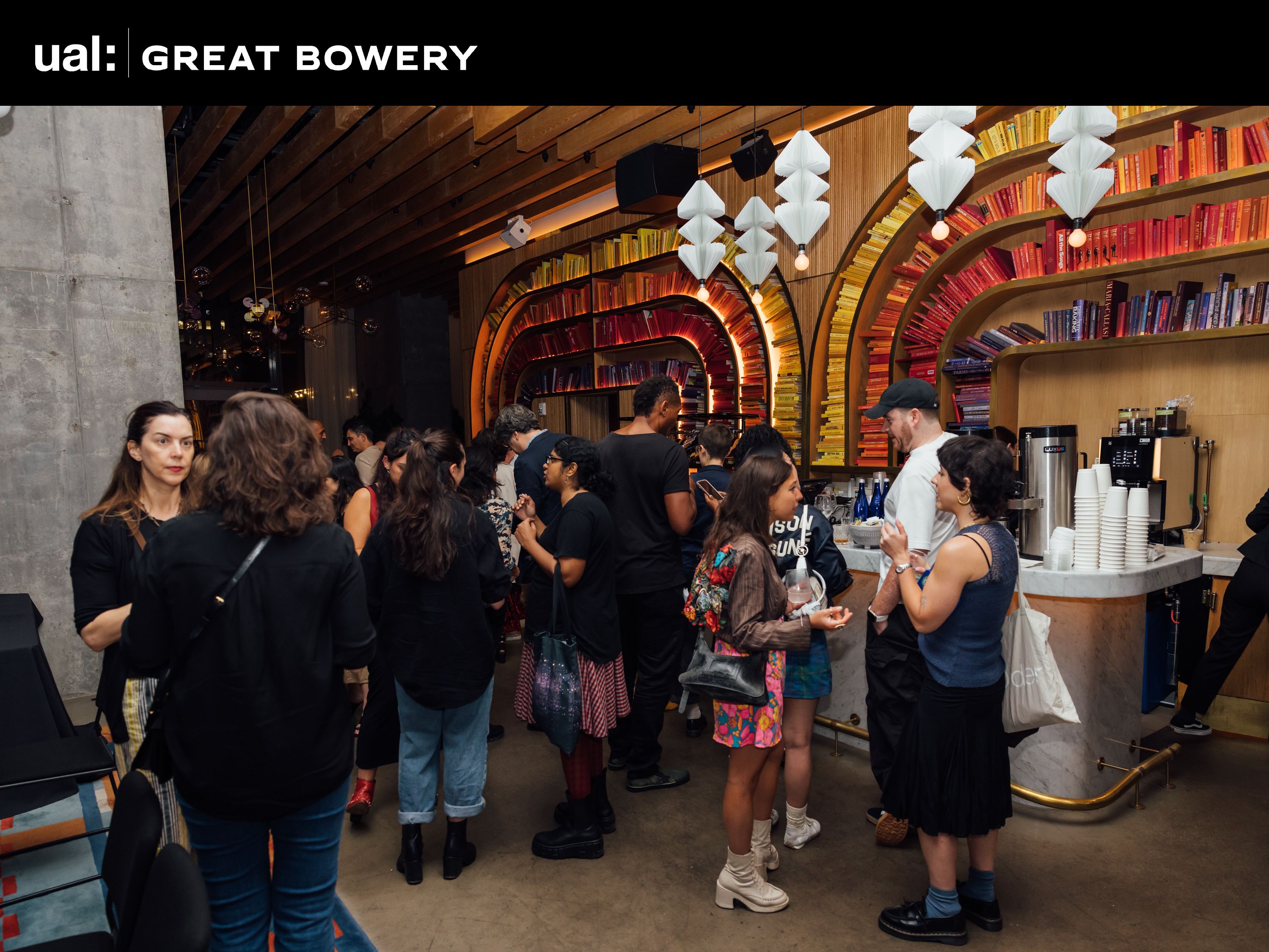 Guests standing at the bar. The wall behind the bar is wood panelled with colour coordinated bookshelves that curve around. 
