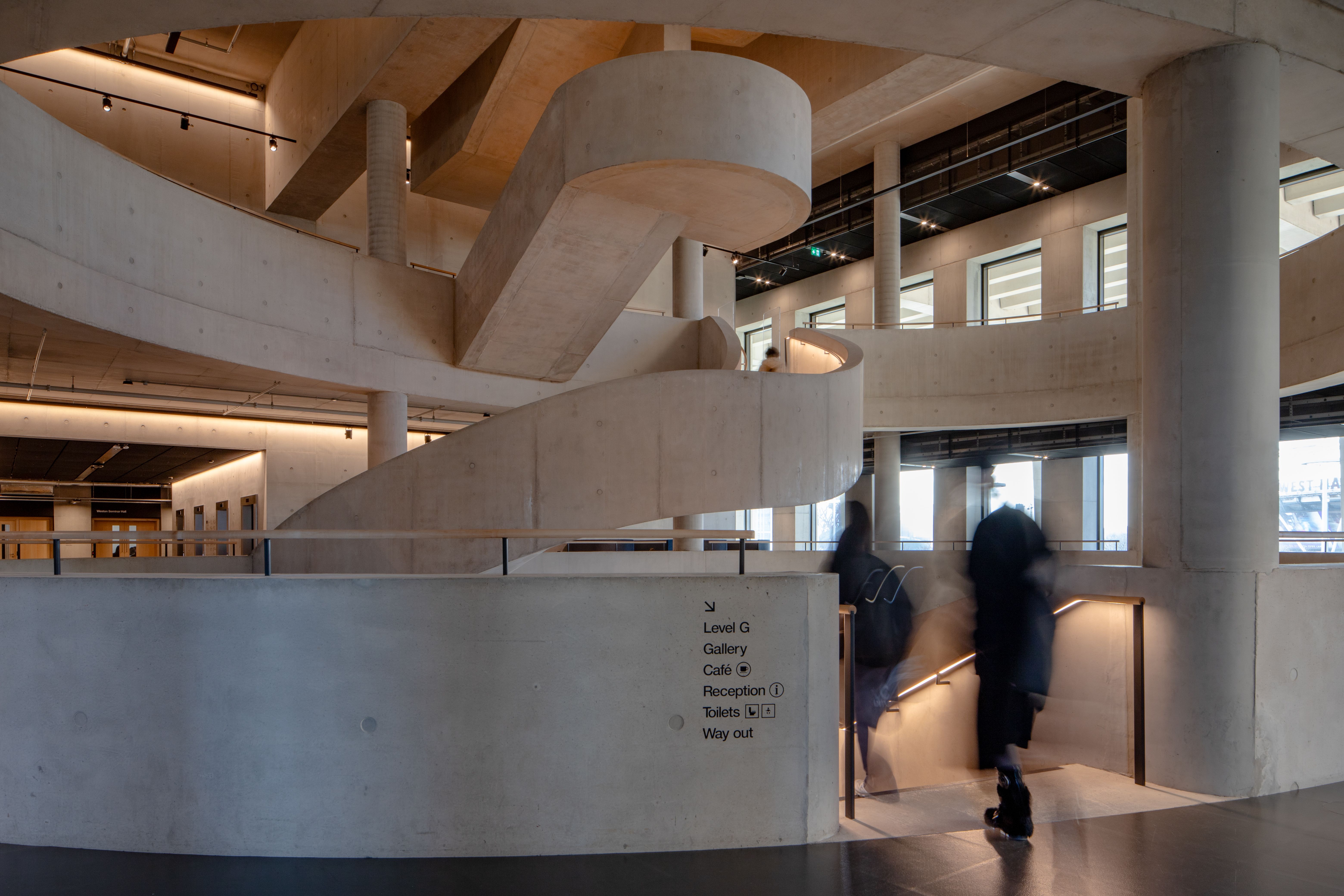Two blurred figures walk down the curved concrete staircase at London College of Fashion.