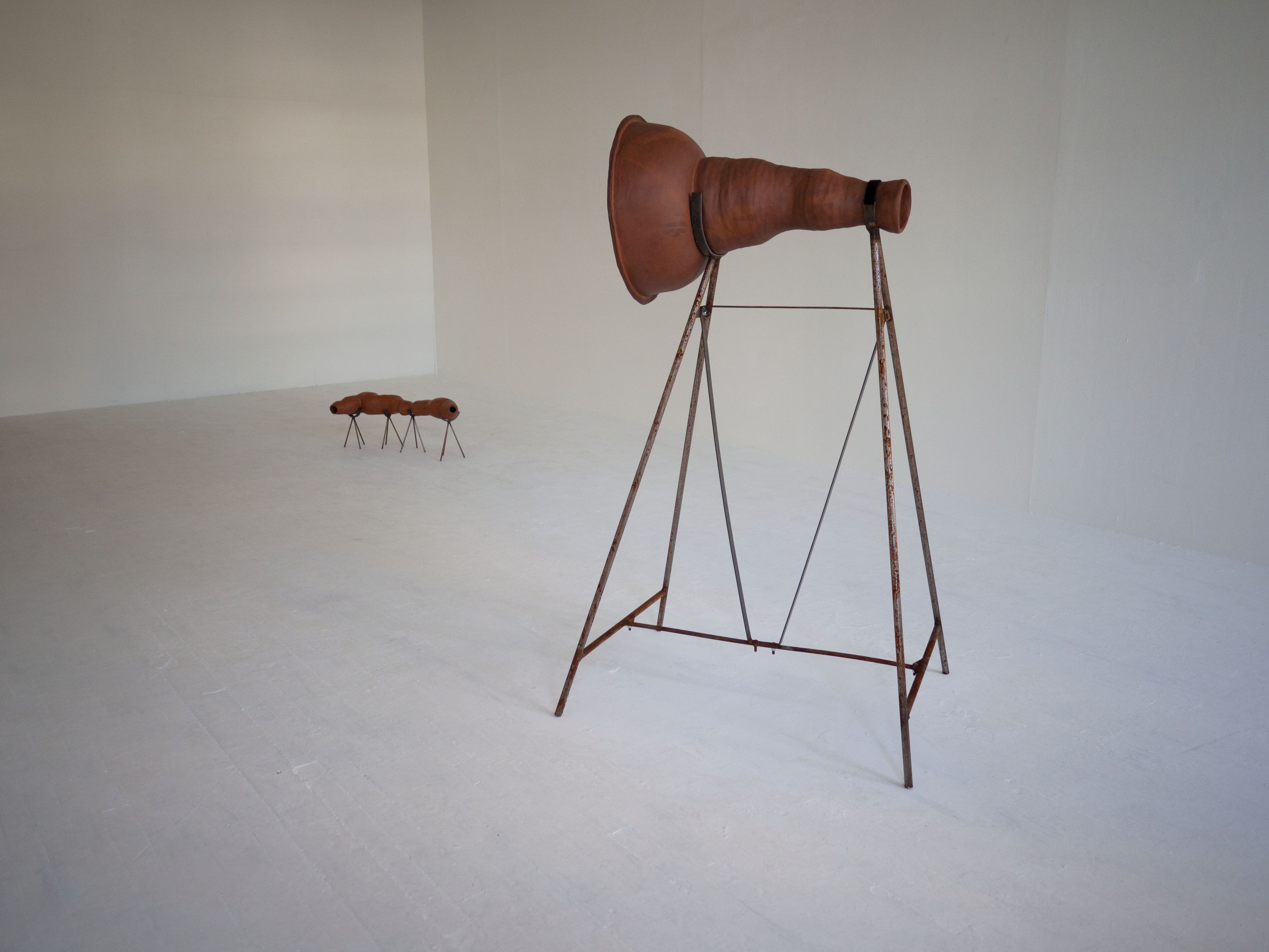 Installation piece, two clay abstract objects are on display in a white walled studio space, both are held up by metal frames.